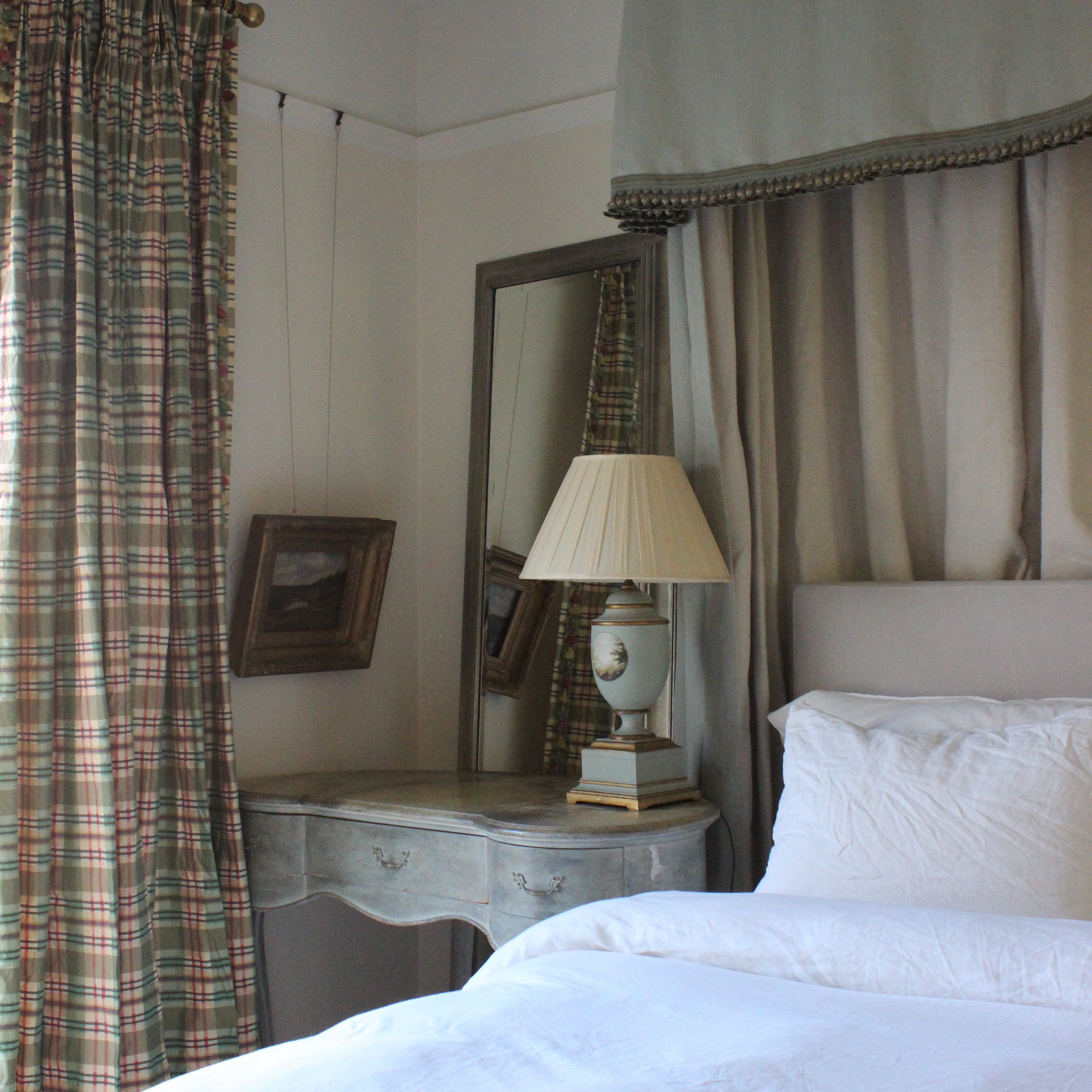 Plaid curtains with English country lamp in blue and gold with mirror and hanging gold frame picture with white sheets on bed with overhead curtains