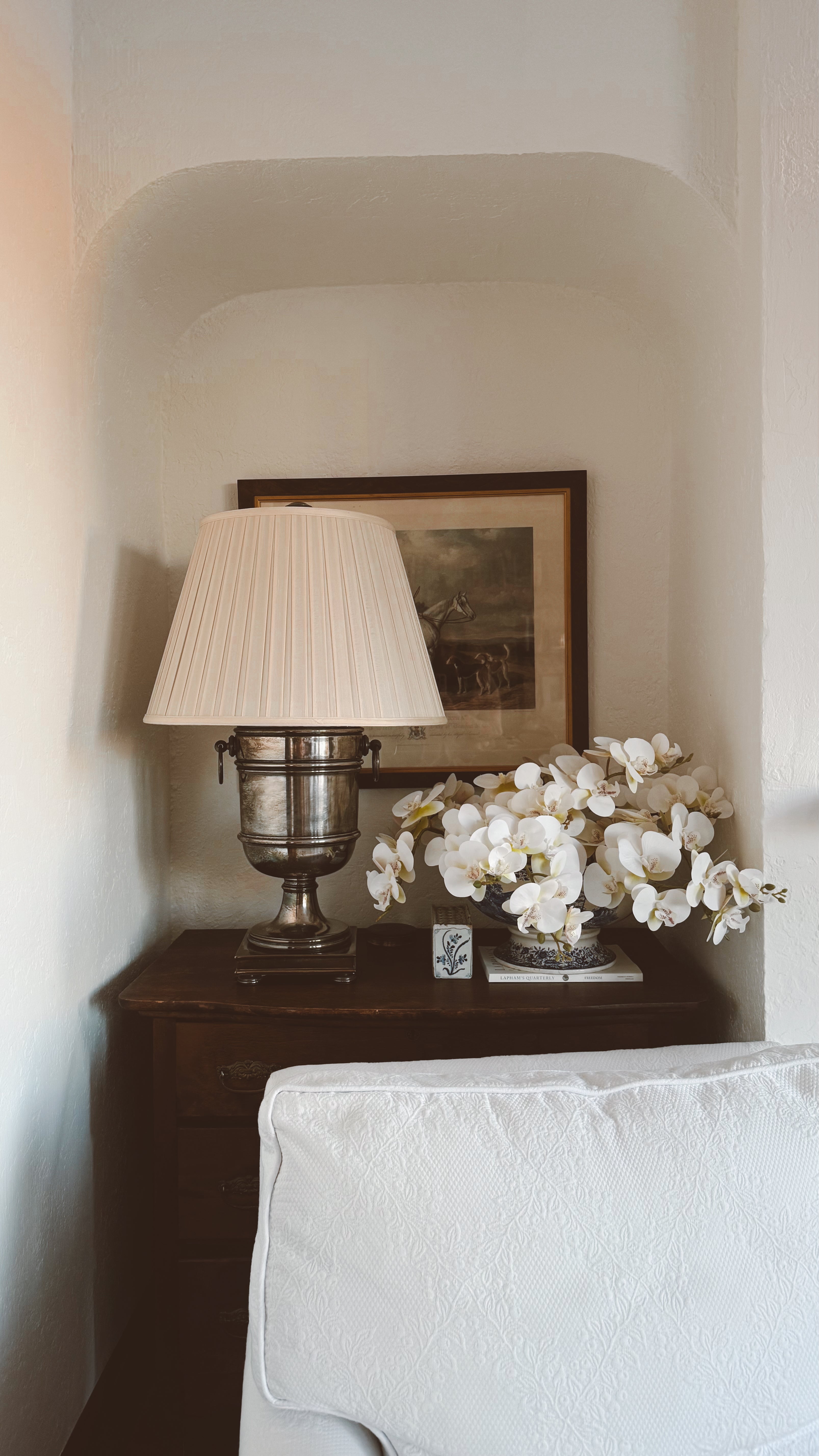 A faux orchid stem bouquet arranged in a antique blue and white bowl in a living room setting, displaying a realistic appearance alongside an antique silver lamp with pleated white shade and a picture frame. Dark wood cabinet and linen sofa.