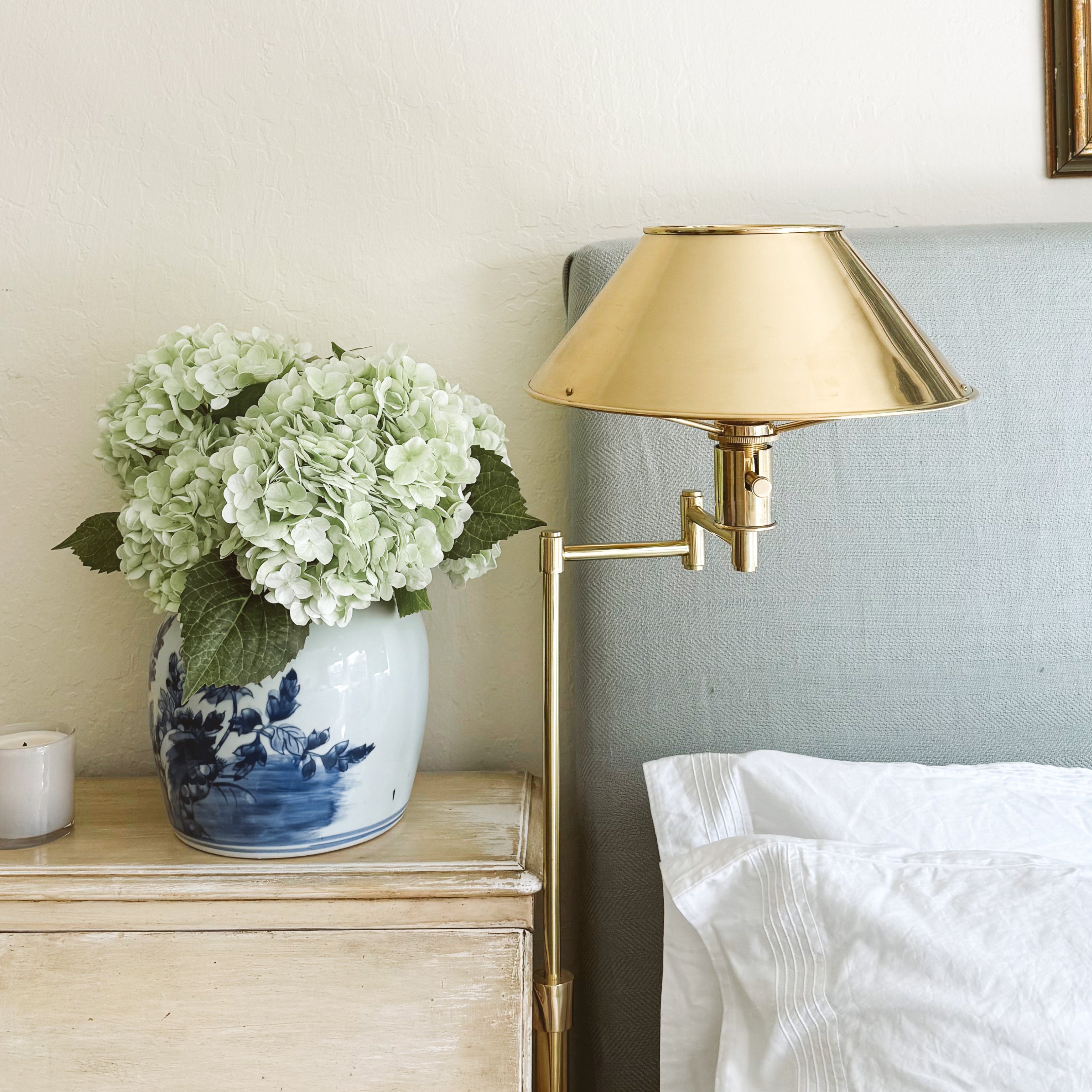 Gold raw brass task lamp with upholstered headboard, white satin pillowcases, candle, and Chinese blue vase with hydrangeas
