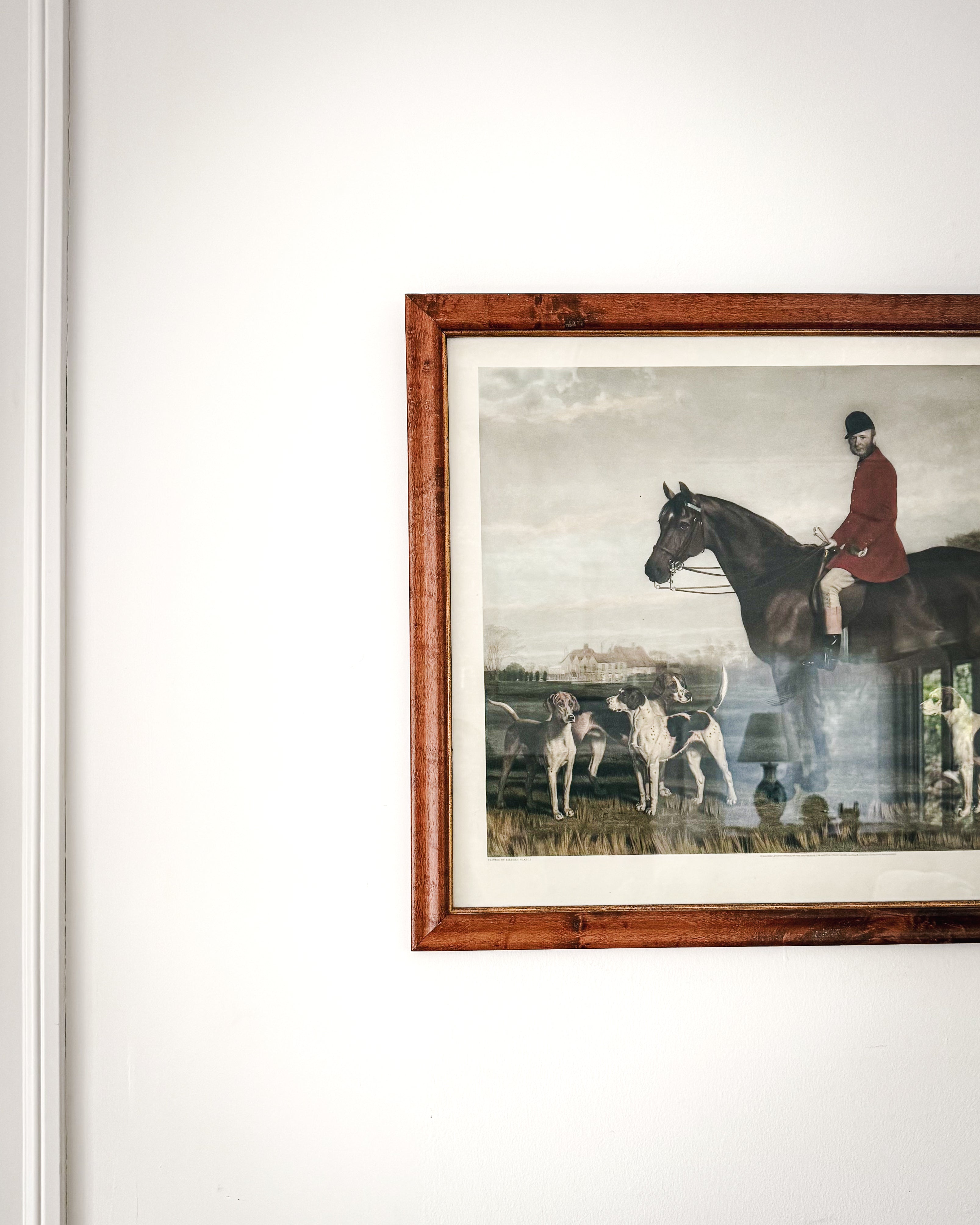 A close up of a framed vintage english art print depicting a classic hunting scene with a rider on horseback and hounds, framed in a burl wood. Hung on a white wall with molding.