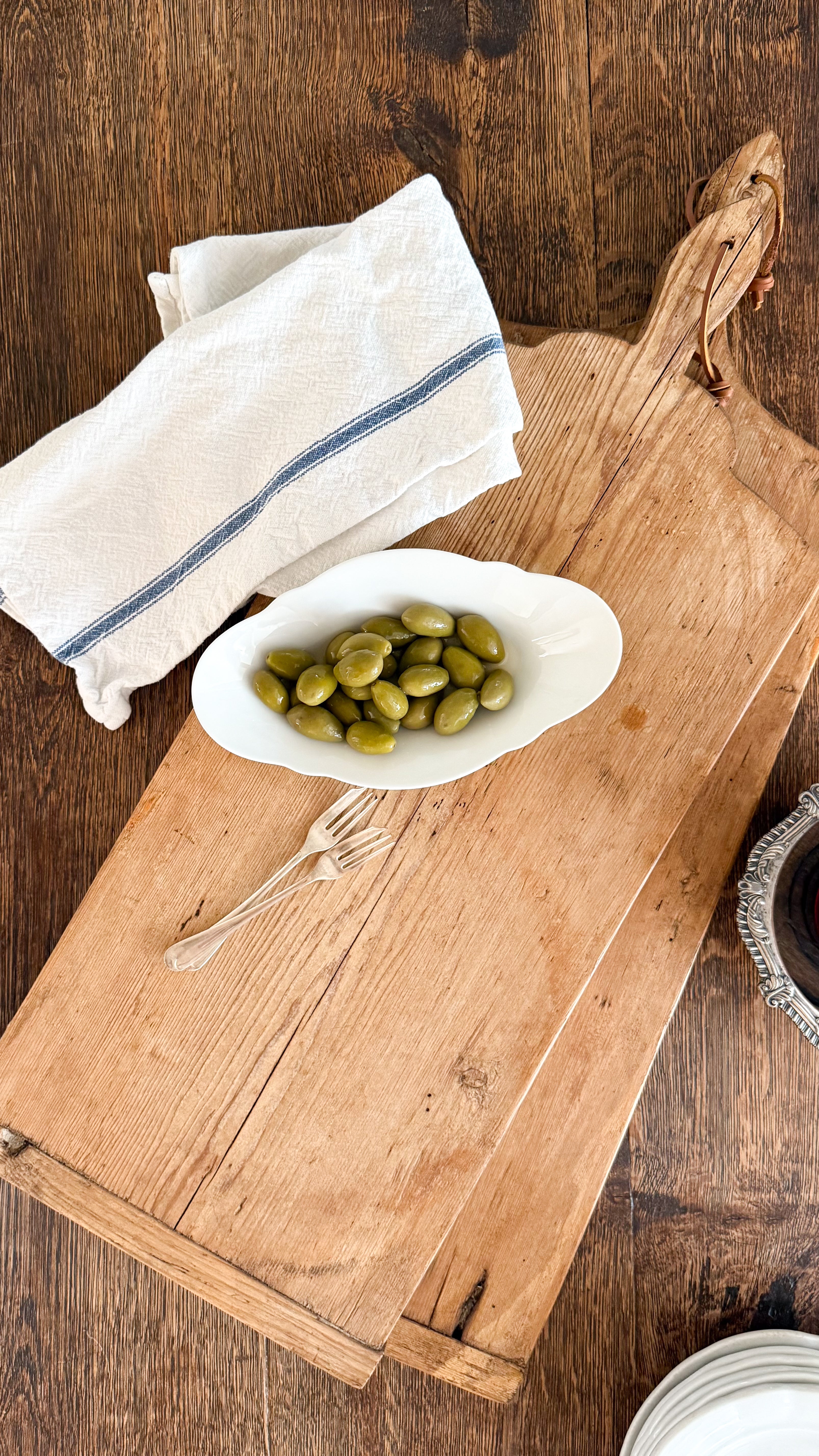 Vintage European Bread board with a bowl of olives, white napkin, and cocktail fork on a Vintage Wooden Dining Table.