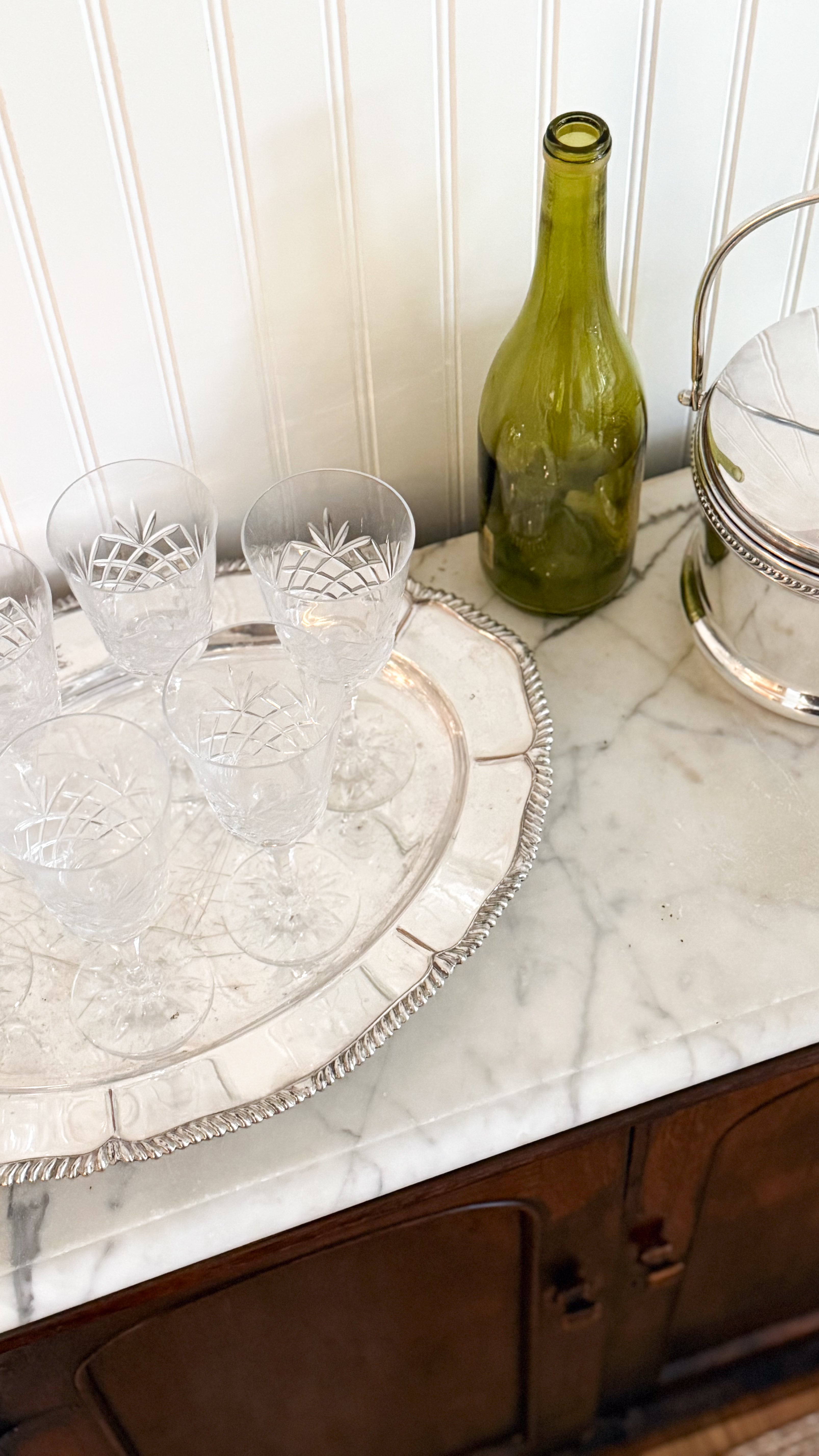 Vintage Irish Cut Crystal Goblets on a Vintage Silver Tray, next to a green glass bottle and a Vintage Silver Ice Bucket; all items on top of a marble sideboard.