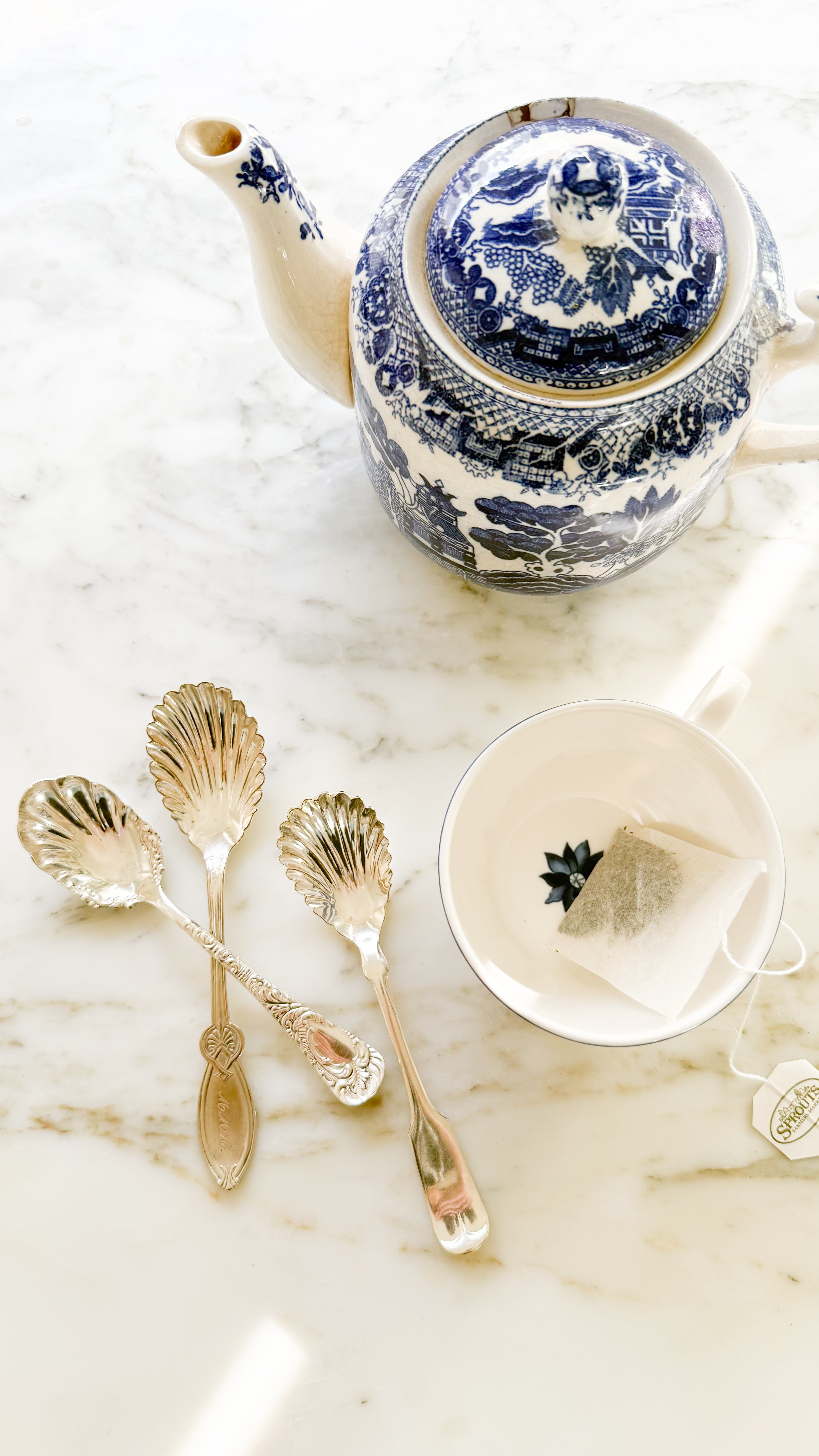 Vintage Silver Sugar and Jelly Spoons on a marble counter with a Vintage blue and white Teapot; a tea cup with a teabag inside