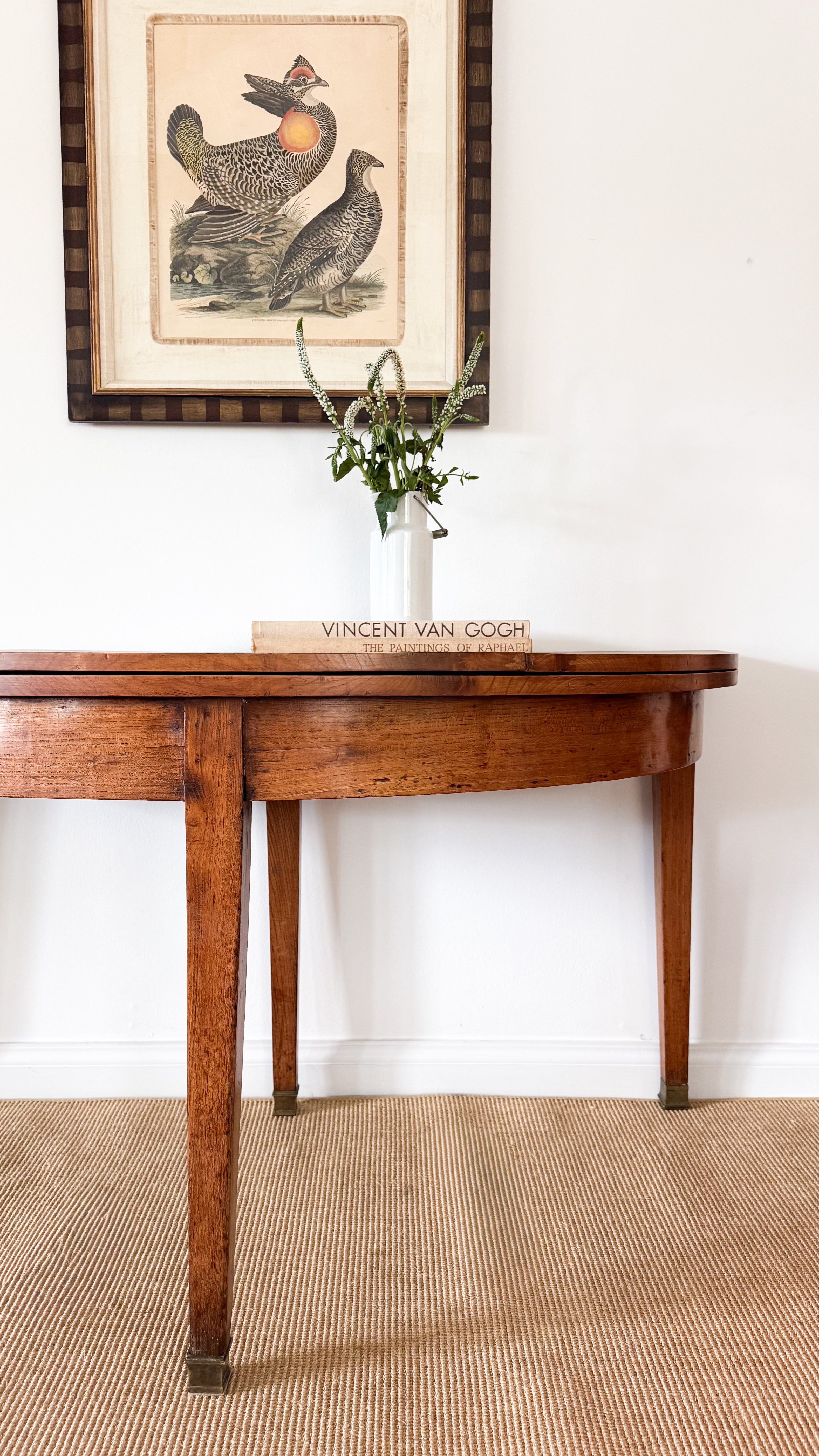Antique wooden dining table with a vase and book, framed artwork above on a white wall.