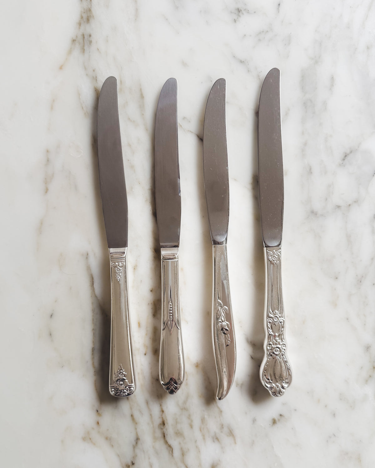 Set of four artfully mismatched vintage silver plate dinner knives with assorted floral and decorative handle patterns, arranged on a white marble surface.