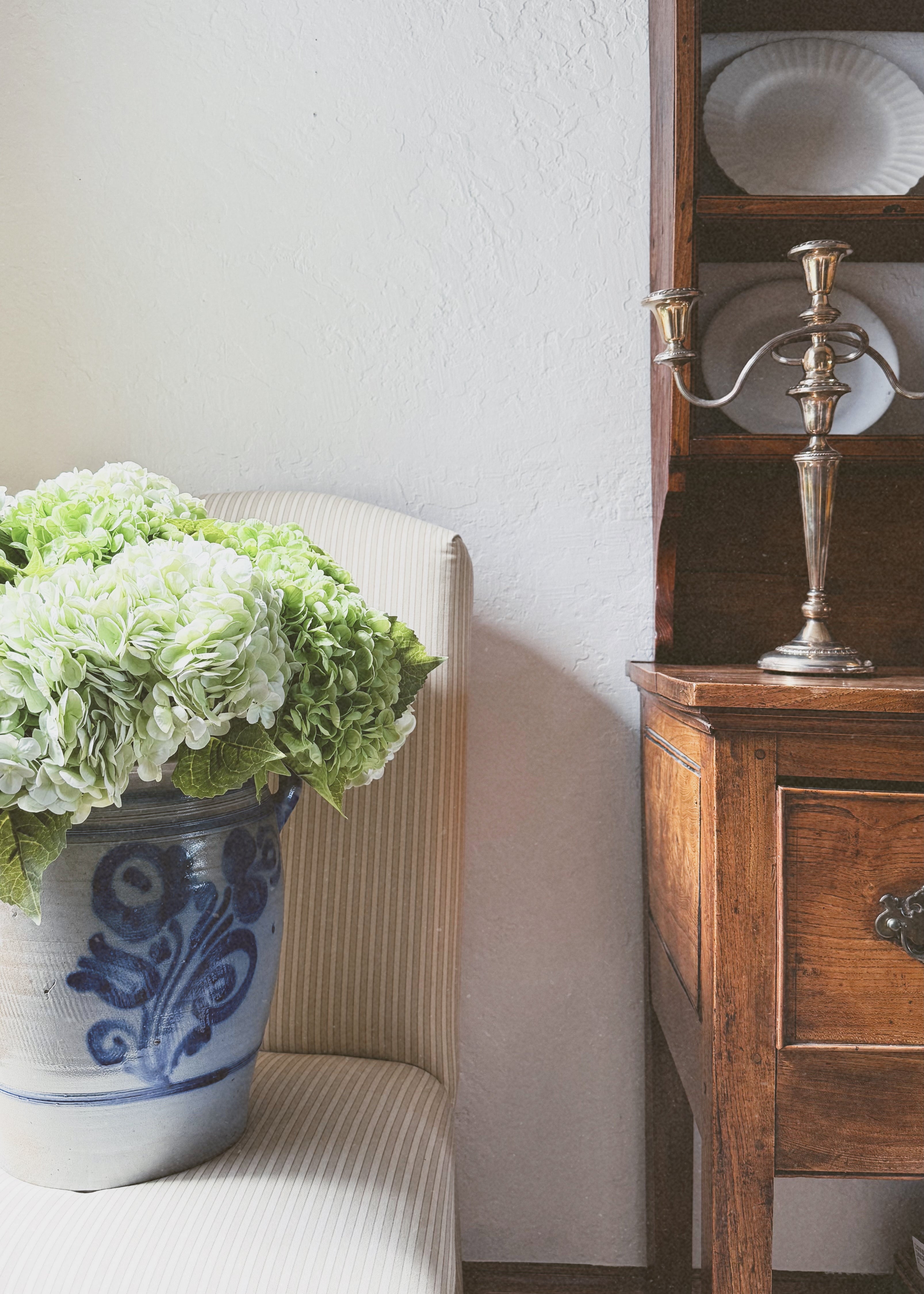 Decorative scene with a vintage crock of hydrangea on a lined upholstered dining chair next to a wooden cabinet with antique silver candleholder