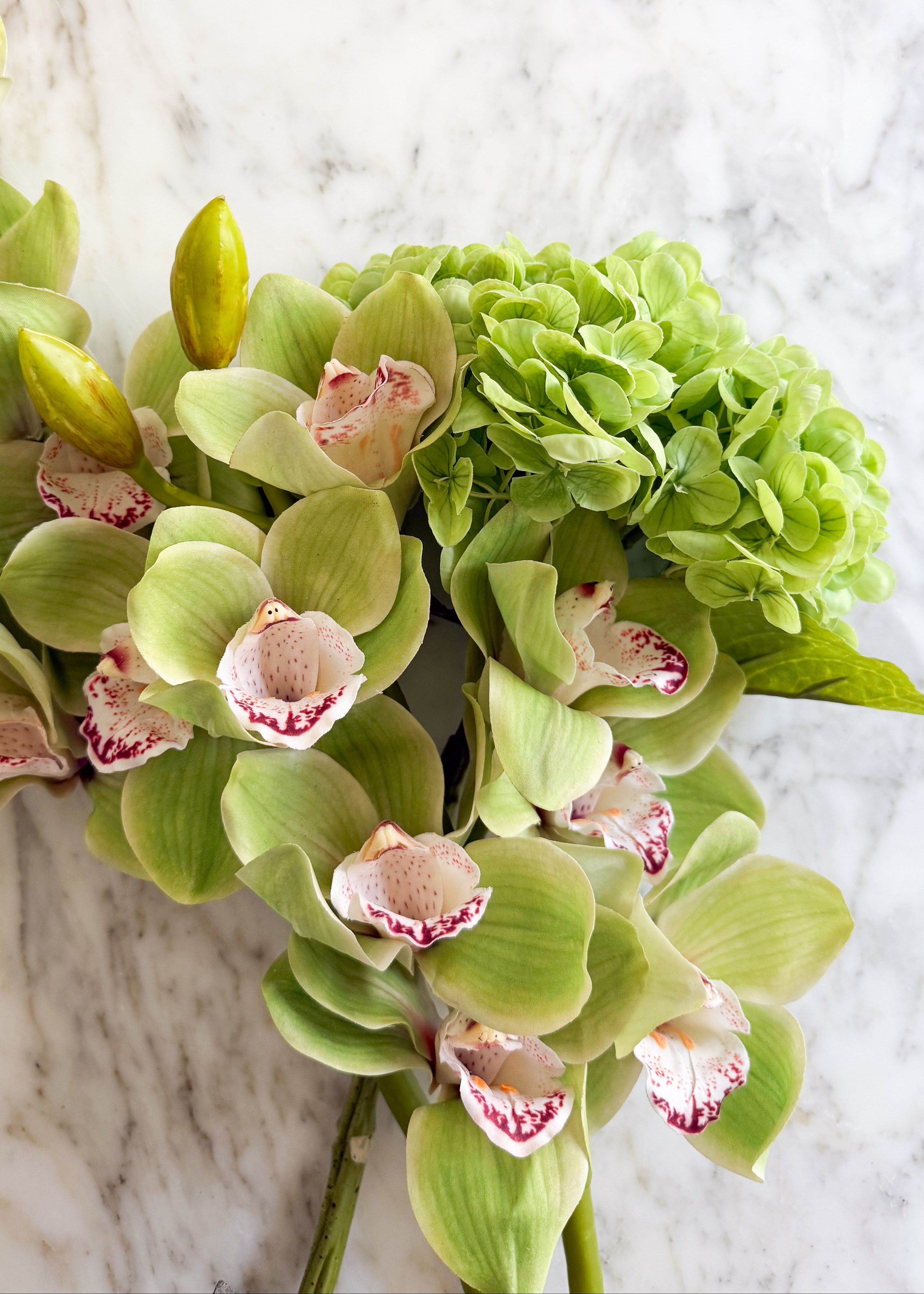 Green faux cymbidium orchid stems paired with a green faux hydrangea stem on a white marble surface.