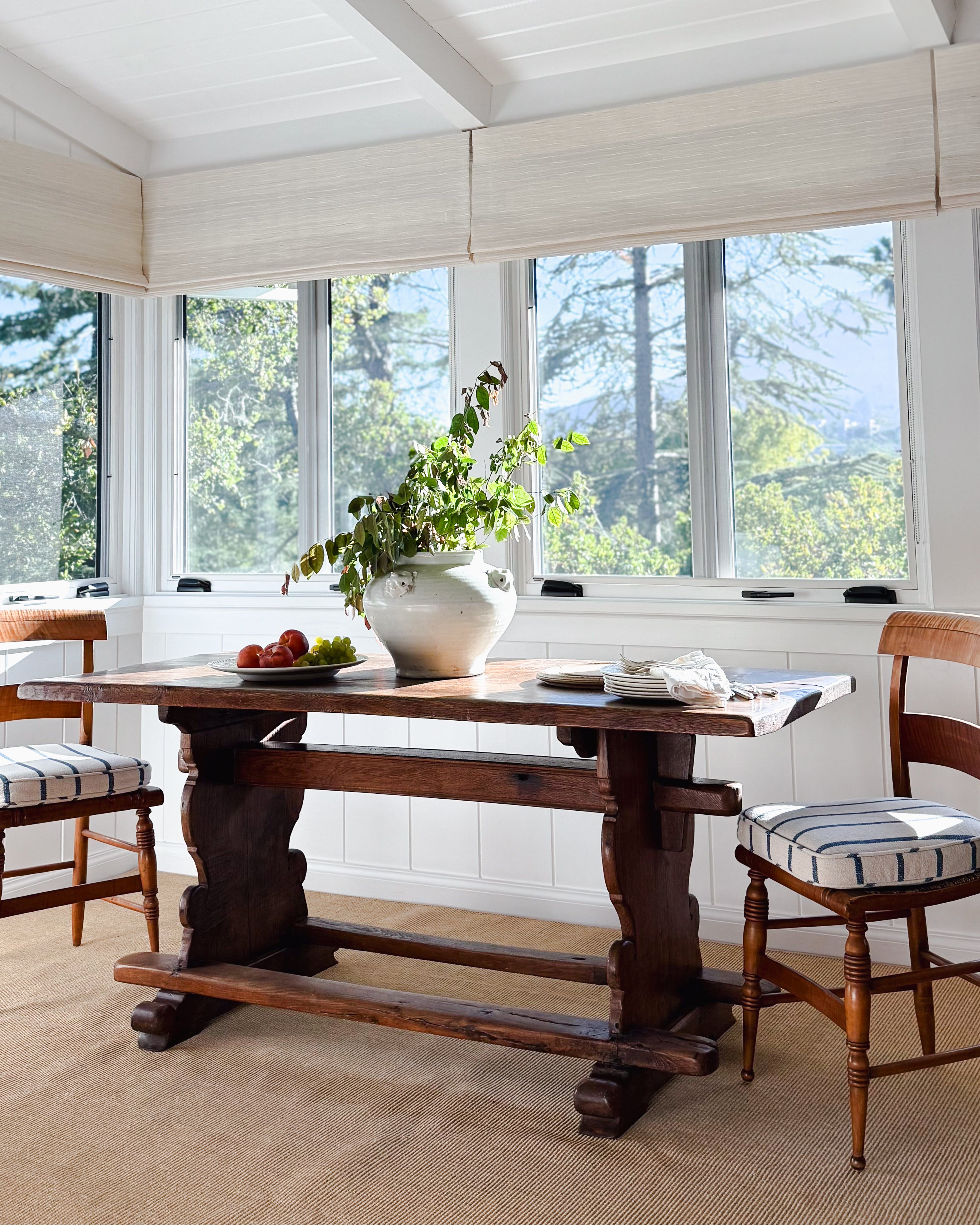 An antique dark wood trestle dining table in a bright, sunlit dining room with large windows, cream walls. Styled with a McConnell handmade estate vessel full of branches.