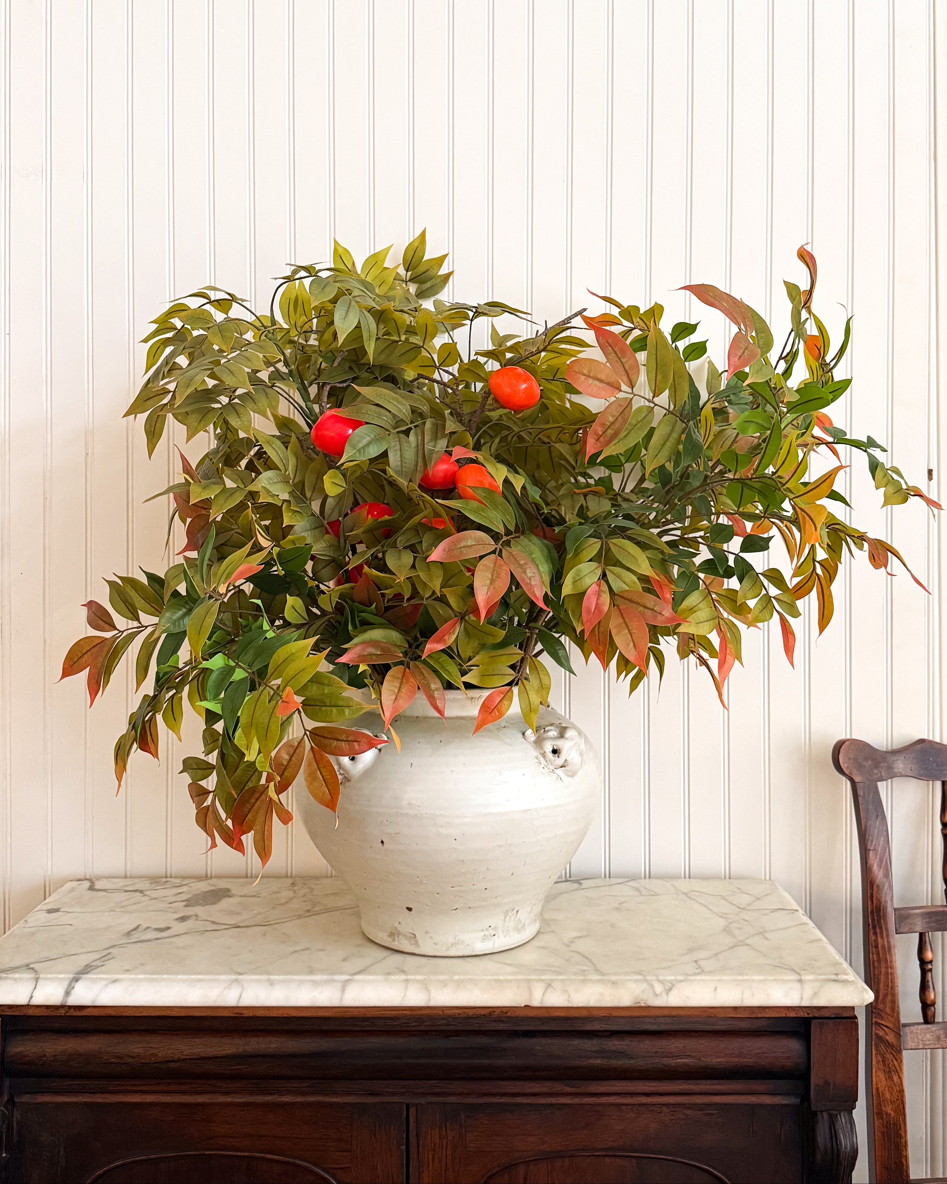 Round Estate Vessel with a handcrafted ivory glaze and lion head accents, styled with faux autumn leaves and persimmon branches on an antique marble-top cabinet.
