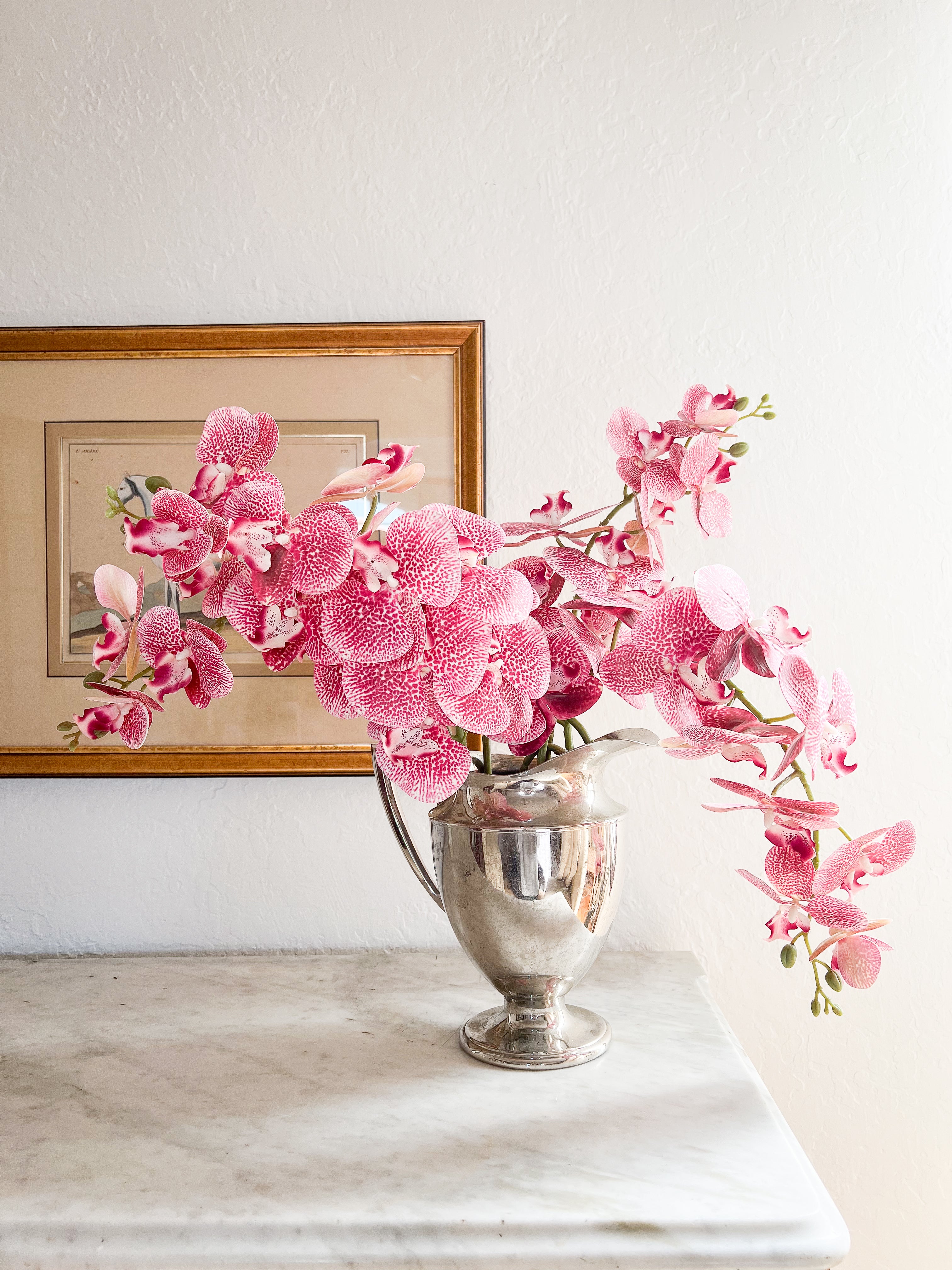 Pink orchids arranged in a vintage silver vase on a marble console table against white wall