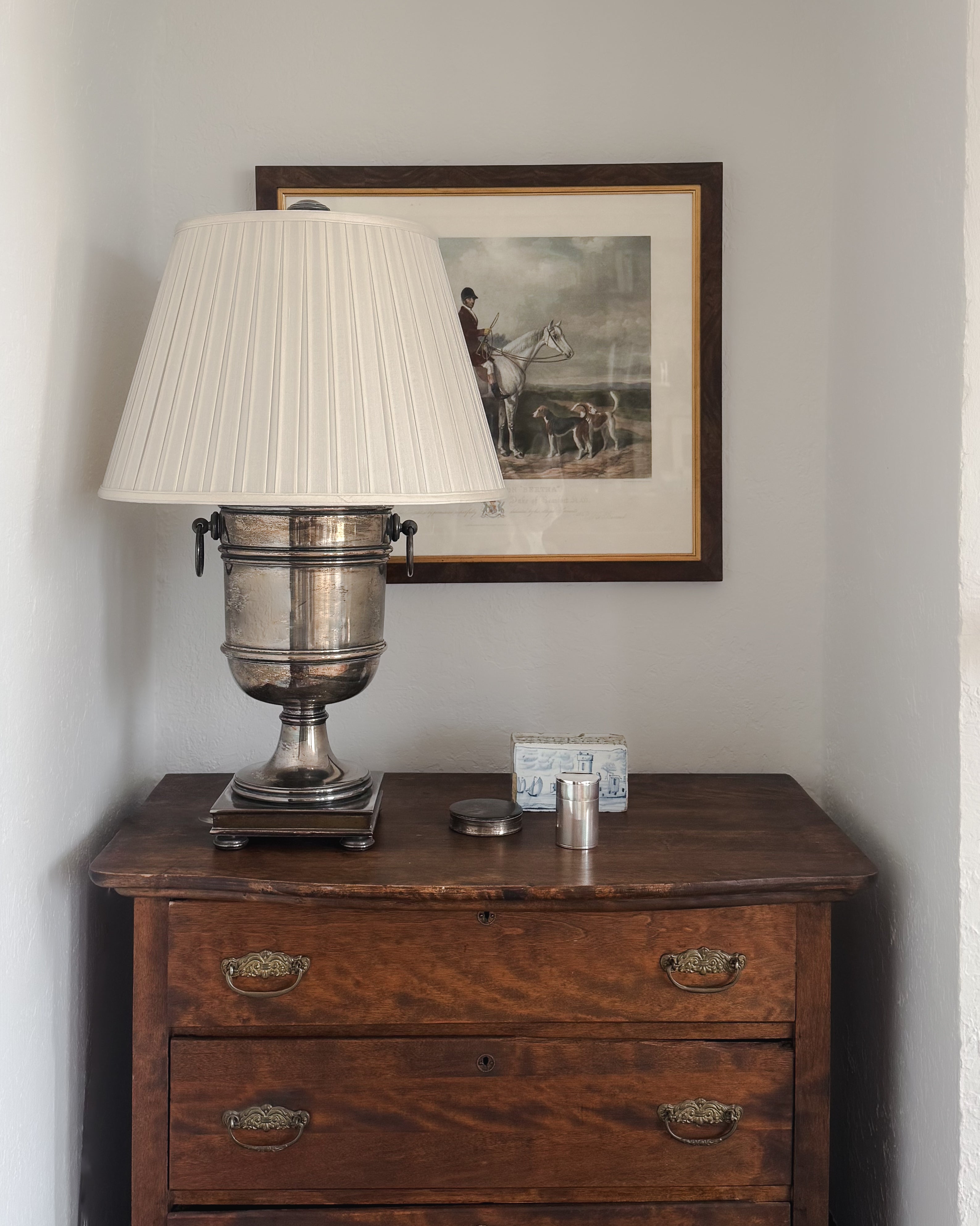 Vintage European silver-plated canister styled on an antique dresser beside a silver urn lamp and framed equestrian print