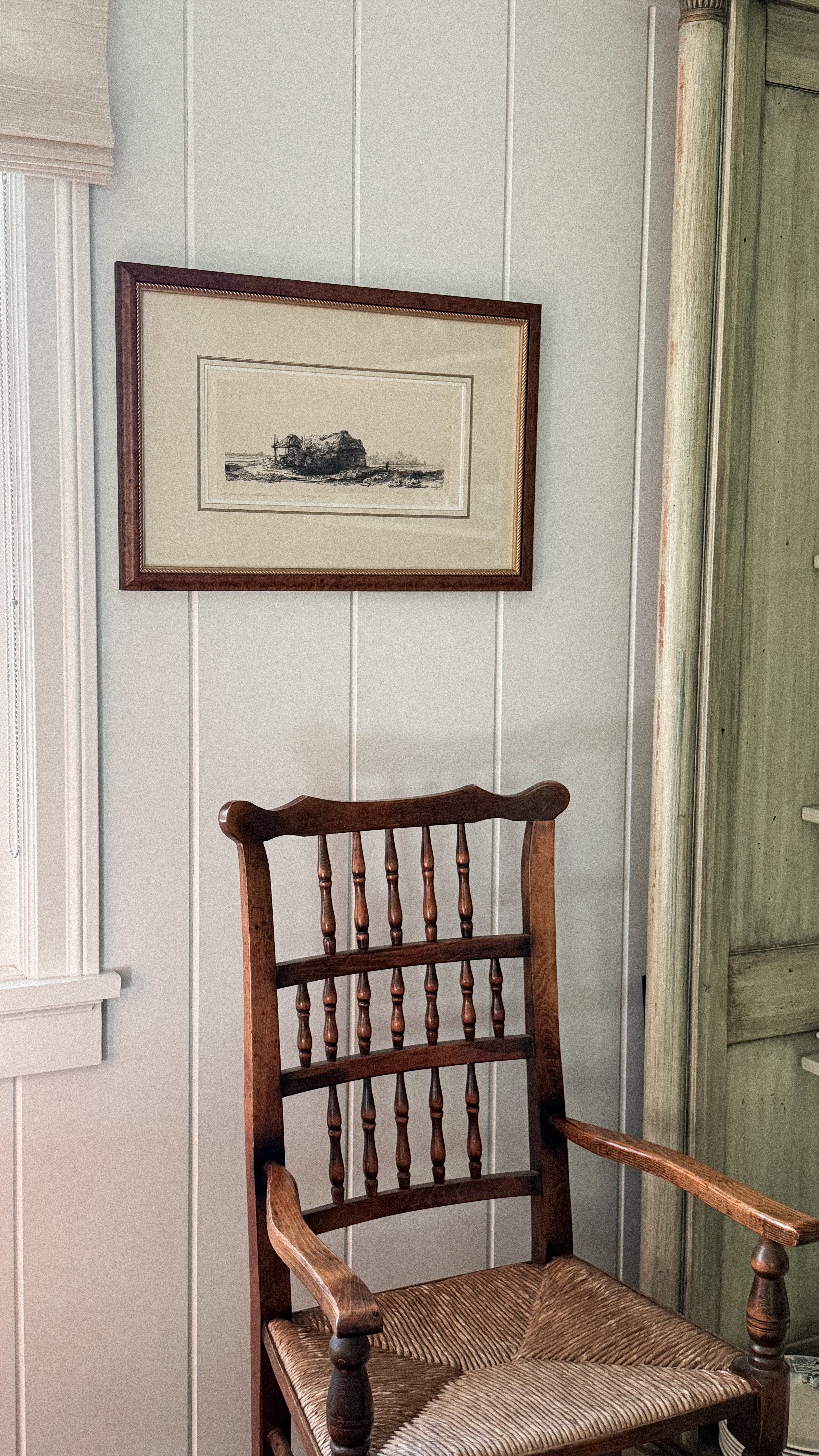 Antique wood chair against a white paneled wall with a framed picture above it.
