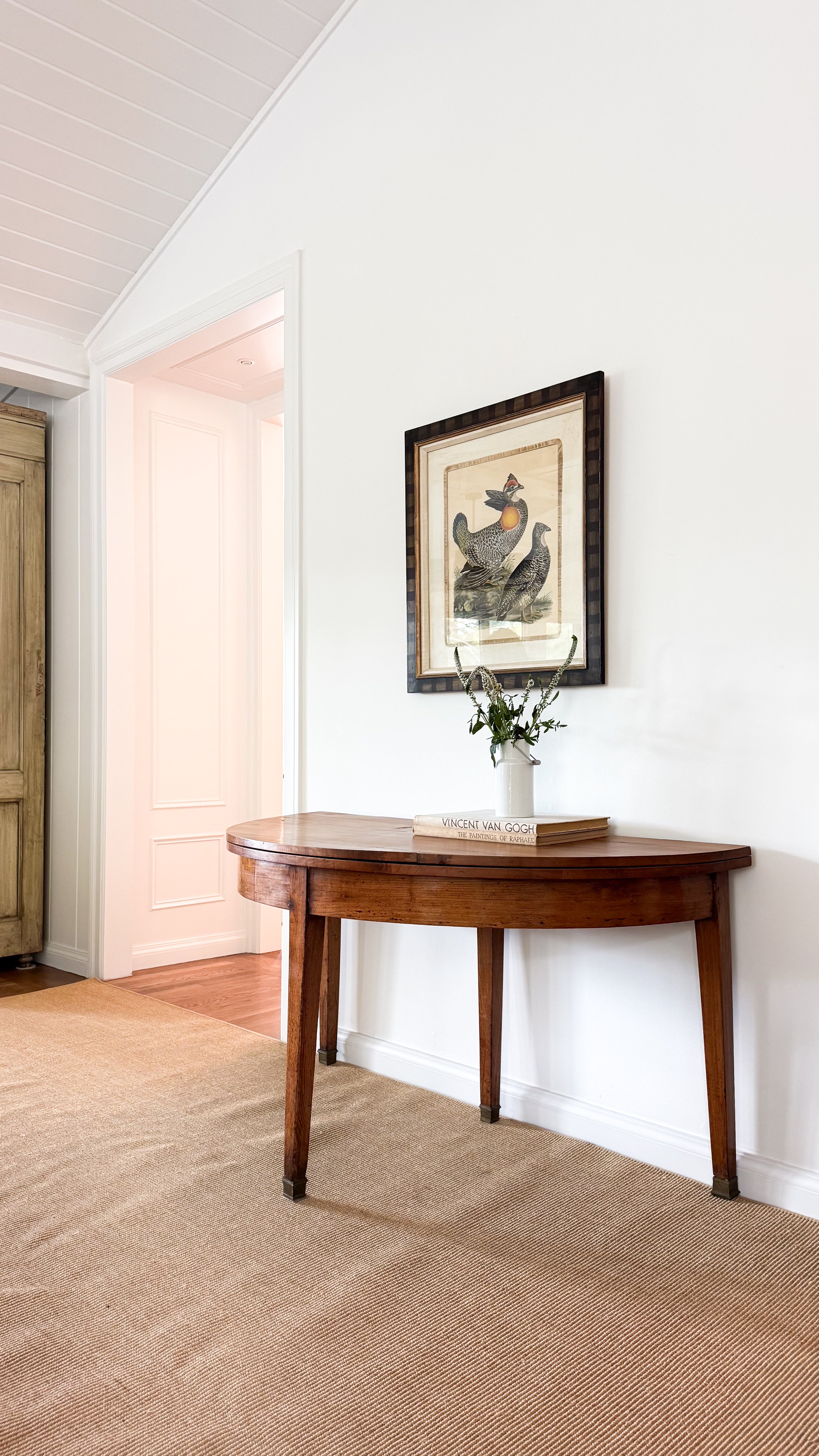 Wooden dining table with a framed picture and plant against a white wall.