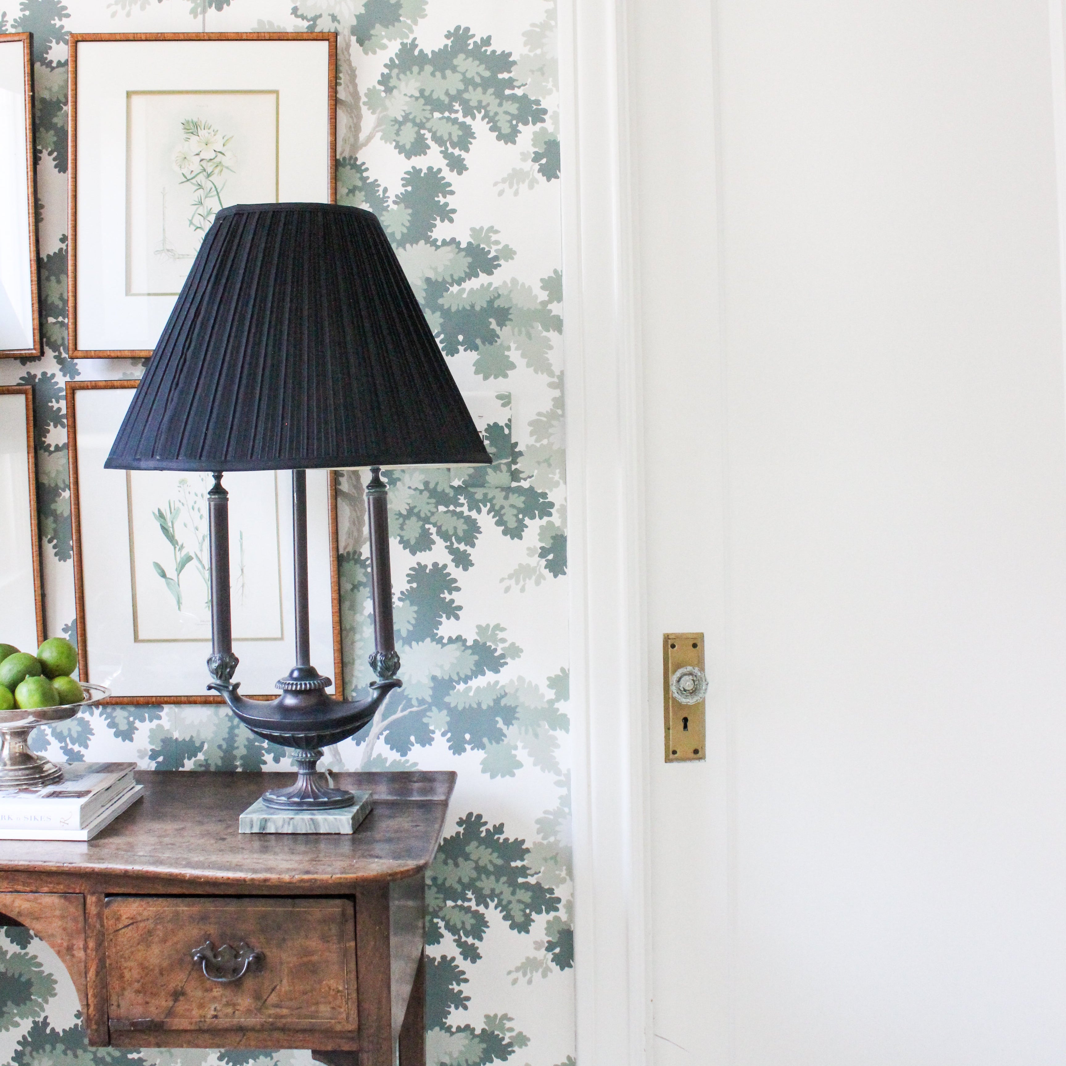 Black lampshade lamp with gold hardware, antique English desk, and green tree wallpaper with gold frame botanical art prints
