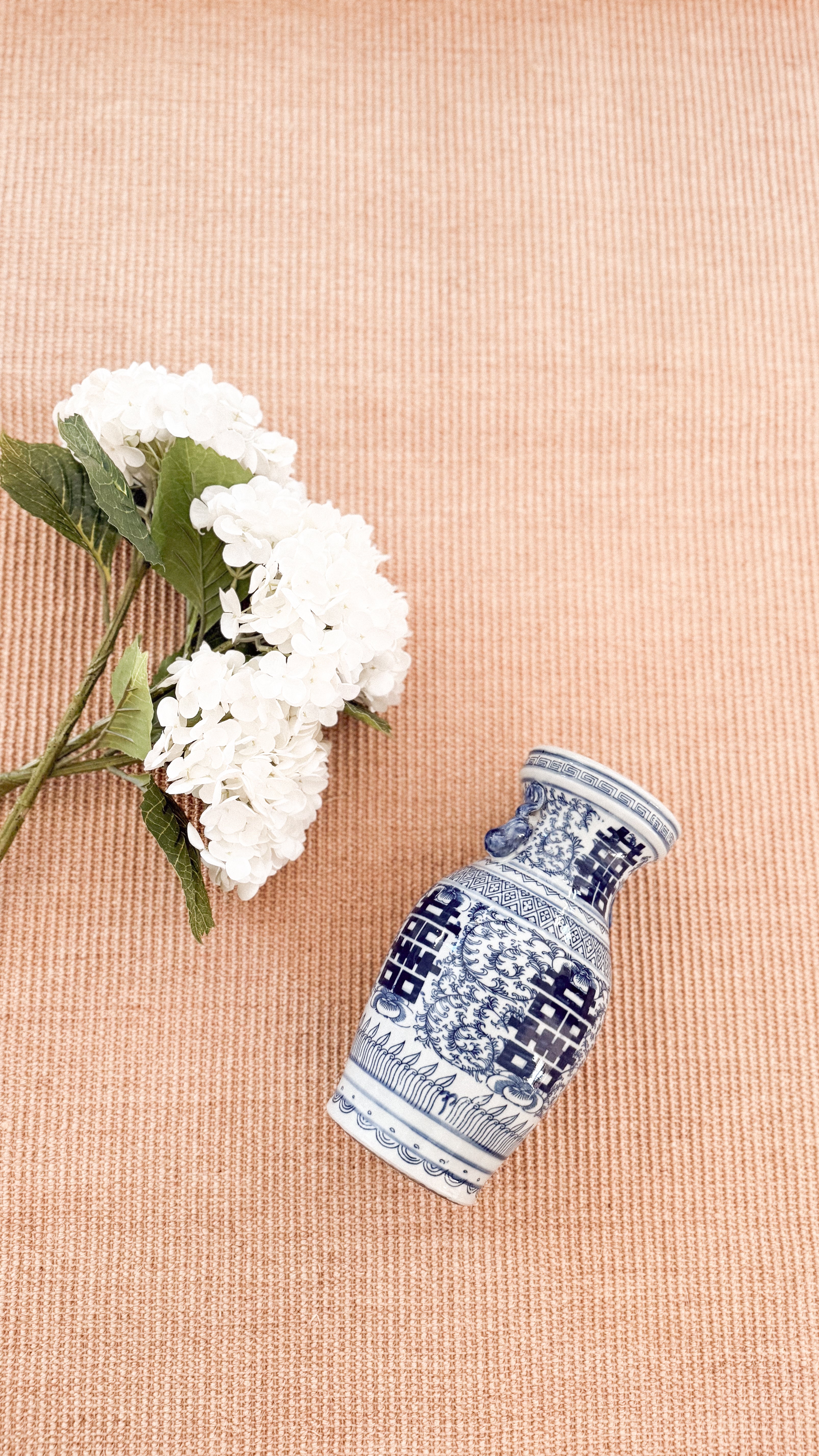 Three Faux White Hydrangeas laying on top of a woven rug, lying alongside a blue and white double happiness vase.
