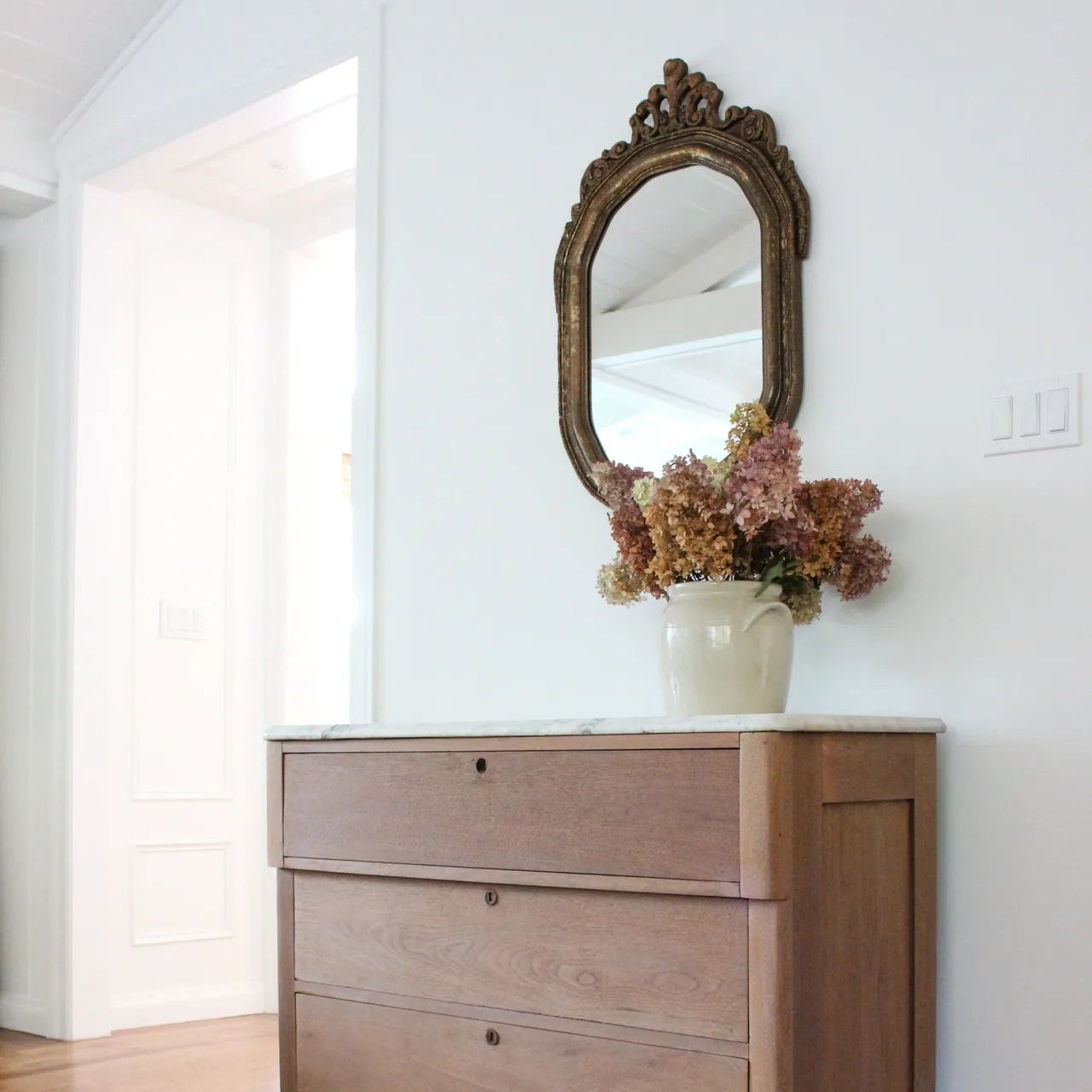 Warm tone solid oak wood cabinet with three drawers and white marble top, white antique vase with florals, and antique mirror with hardwood floors and white walls.