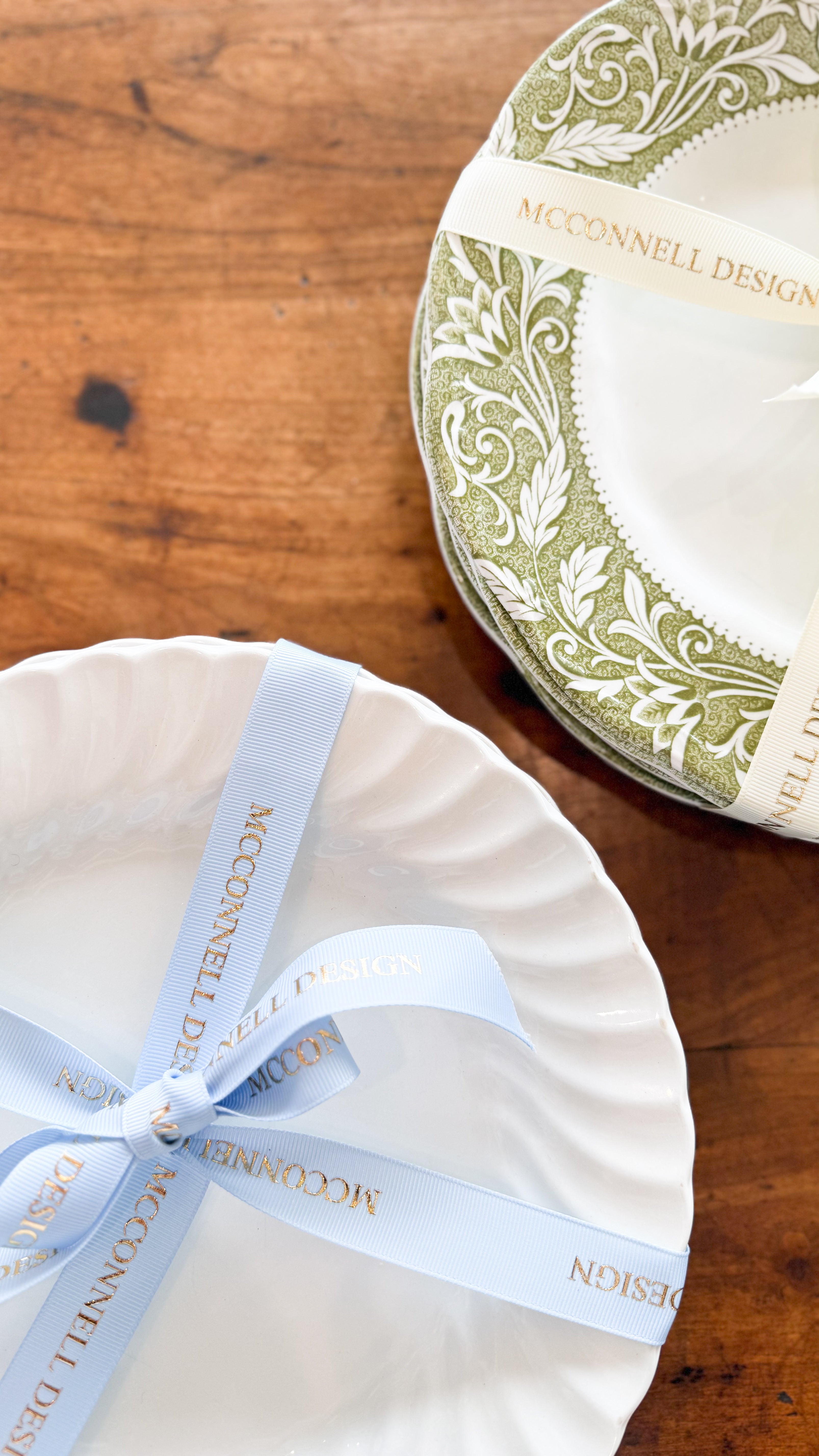  Set of 6 White Scalloped dishes, tied together with a blue McConnell Design ribbon, next to another Set of 6 Green Border Dishes; tied with a ivory McConnell Design ribbon, on top of a vintage table.