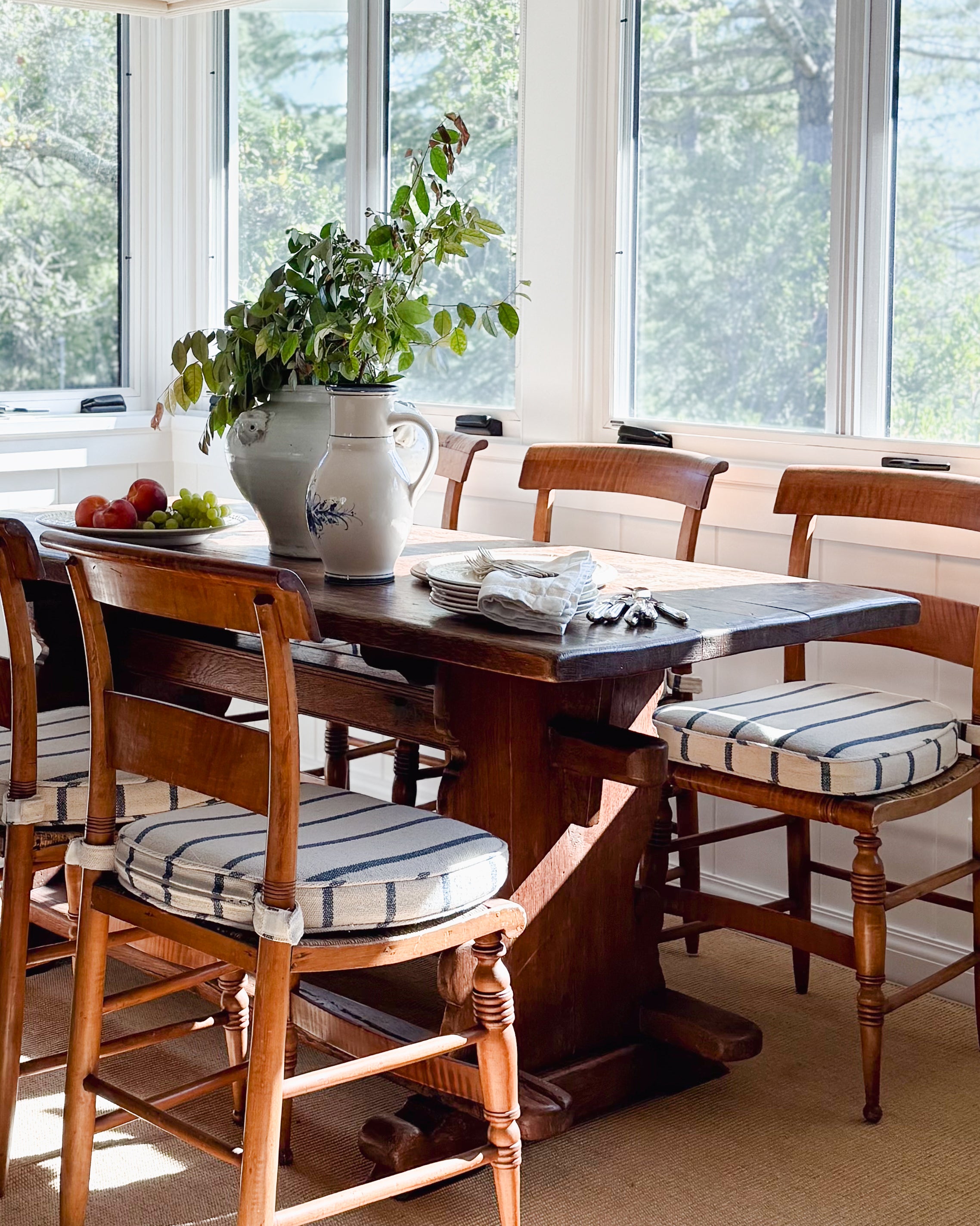 A vintage stoneware pitcher pictured on an antique wood trestle dining table with antique wood chairs featuring blue and white striped seat cushions, set in a bright sunroom with large windows.