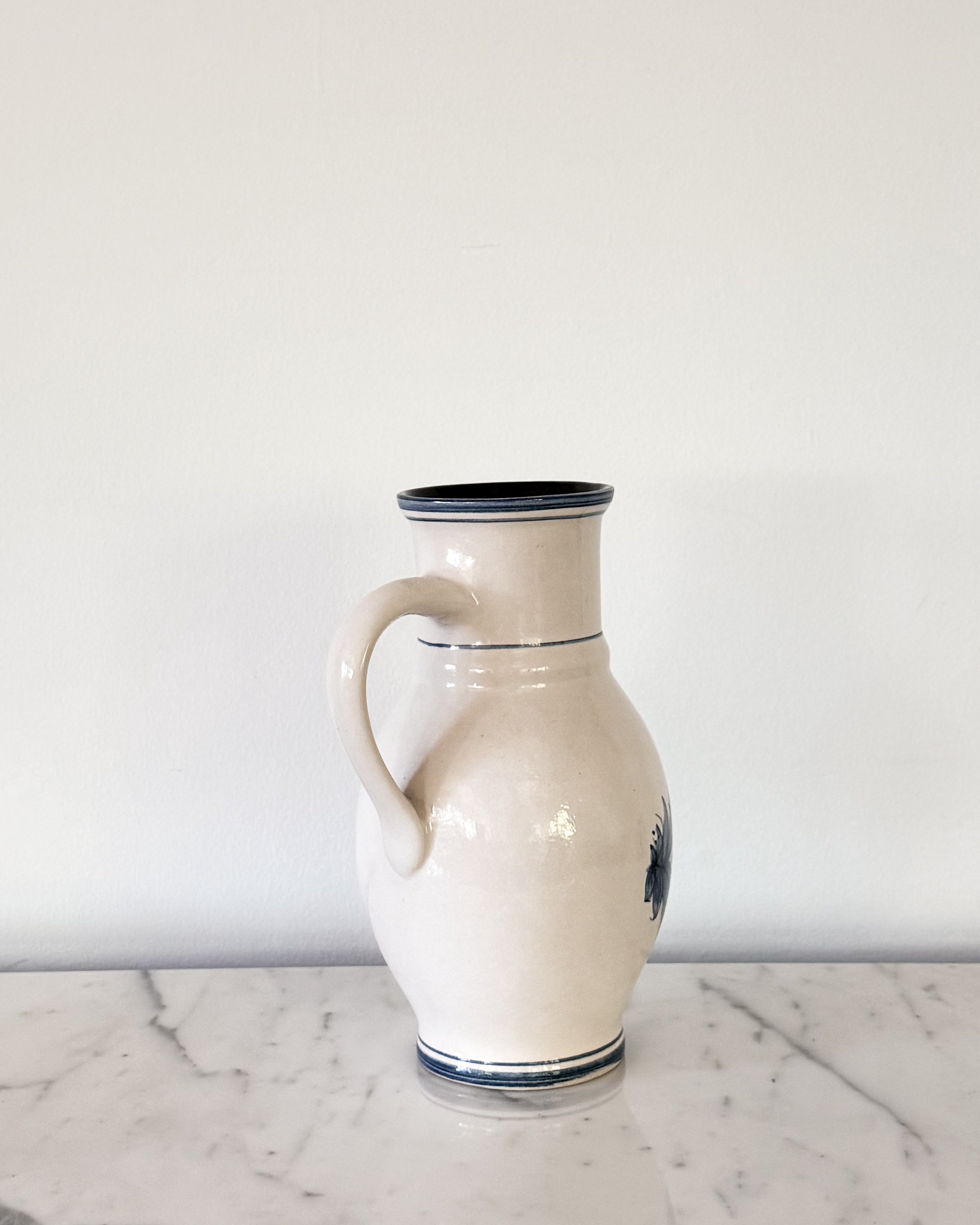 A vintage off-white ceramic pitcher with a dark blue rim and hand painted blue decorative accents, displayed on a white marble surface against a white wall.