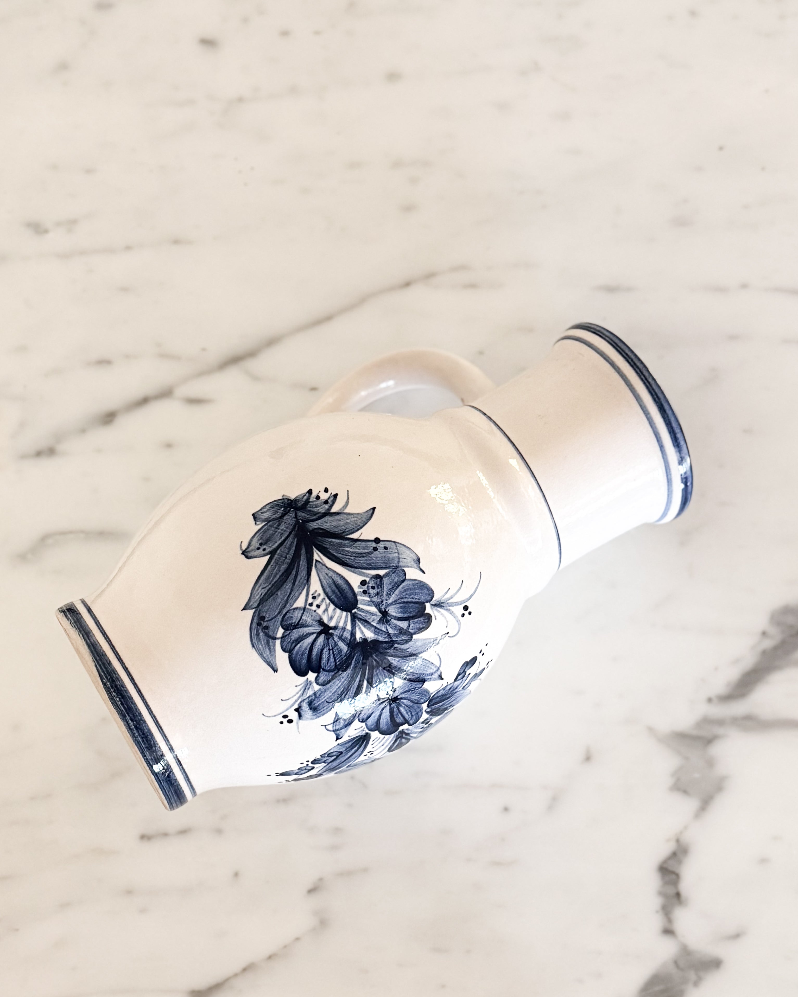 A vintage white ceramic pitcher with a hand painted blue floral pattern and blue rim detailing, photographed on a white marble surface.