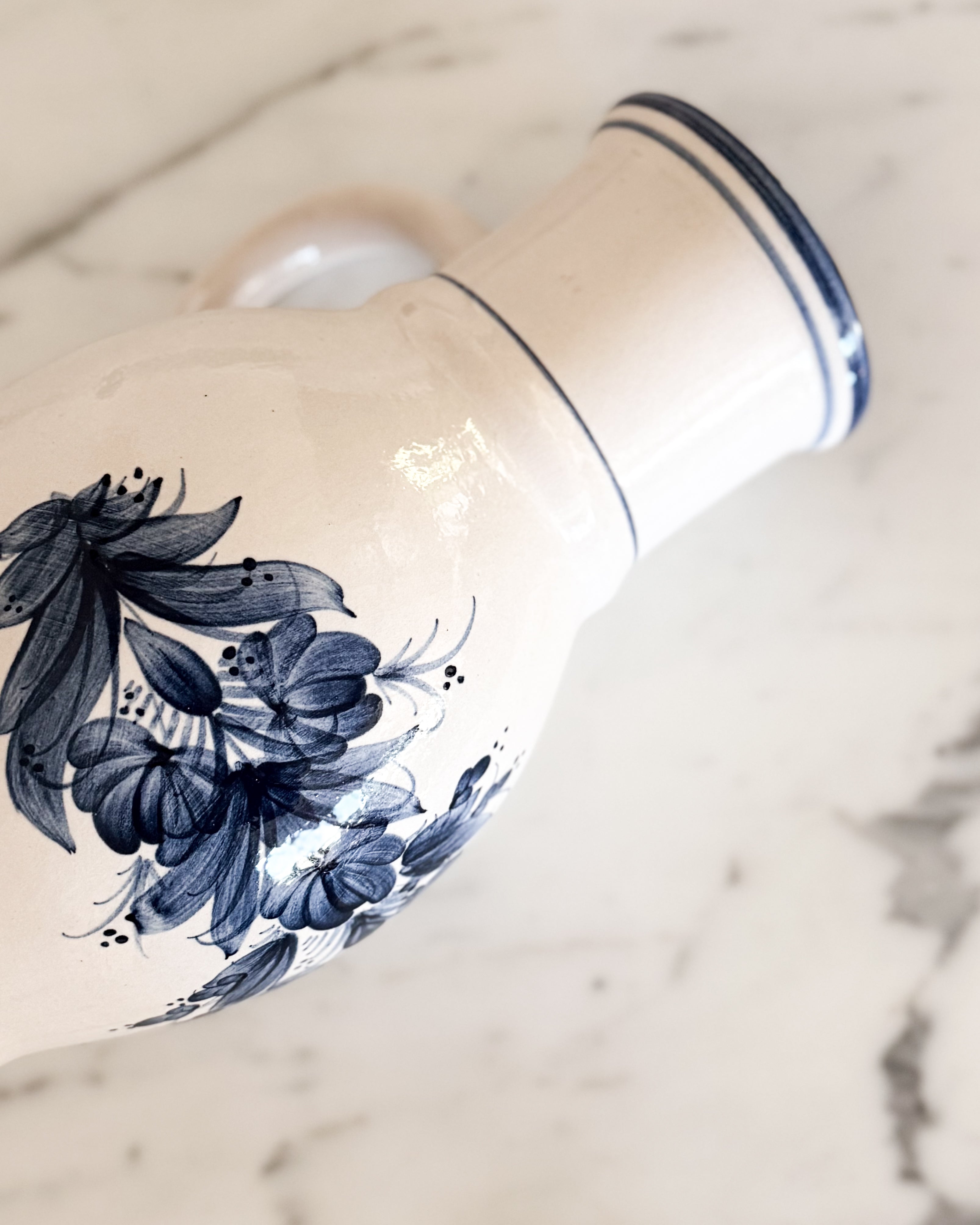 Close up of a vintage creamy white vintage pitcher with hand painted floral details and a painted blue rim on top of a marble surface. 