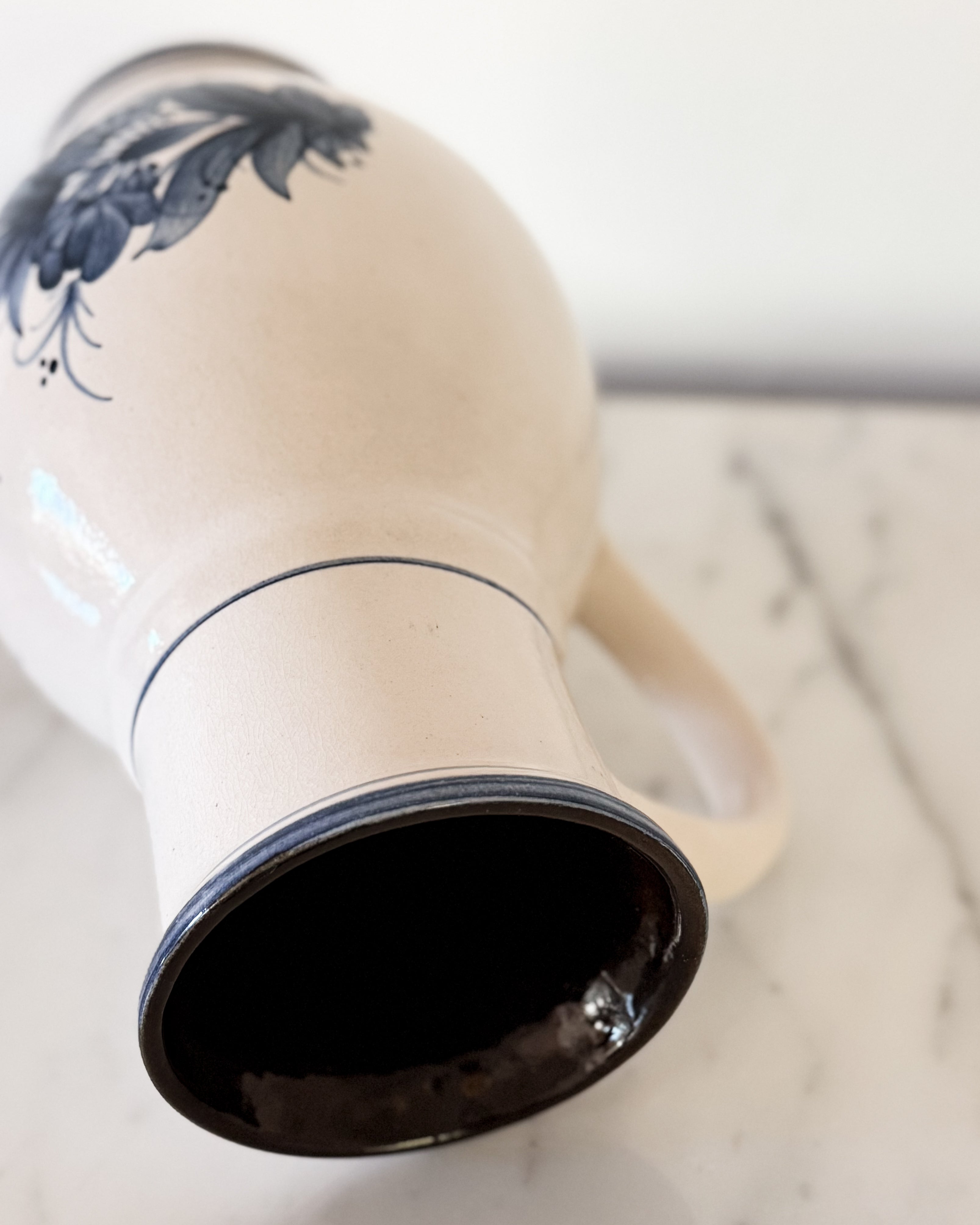 A close-up of a vintage cream-colored ceramic pitcher with a dark blue floral pattern and a dark glazed interior, resting on a white marble surface.