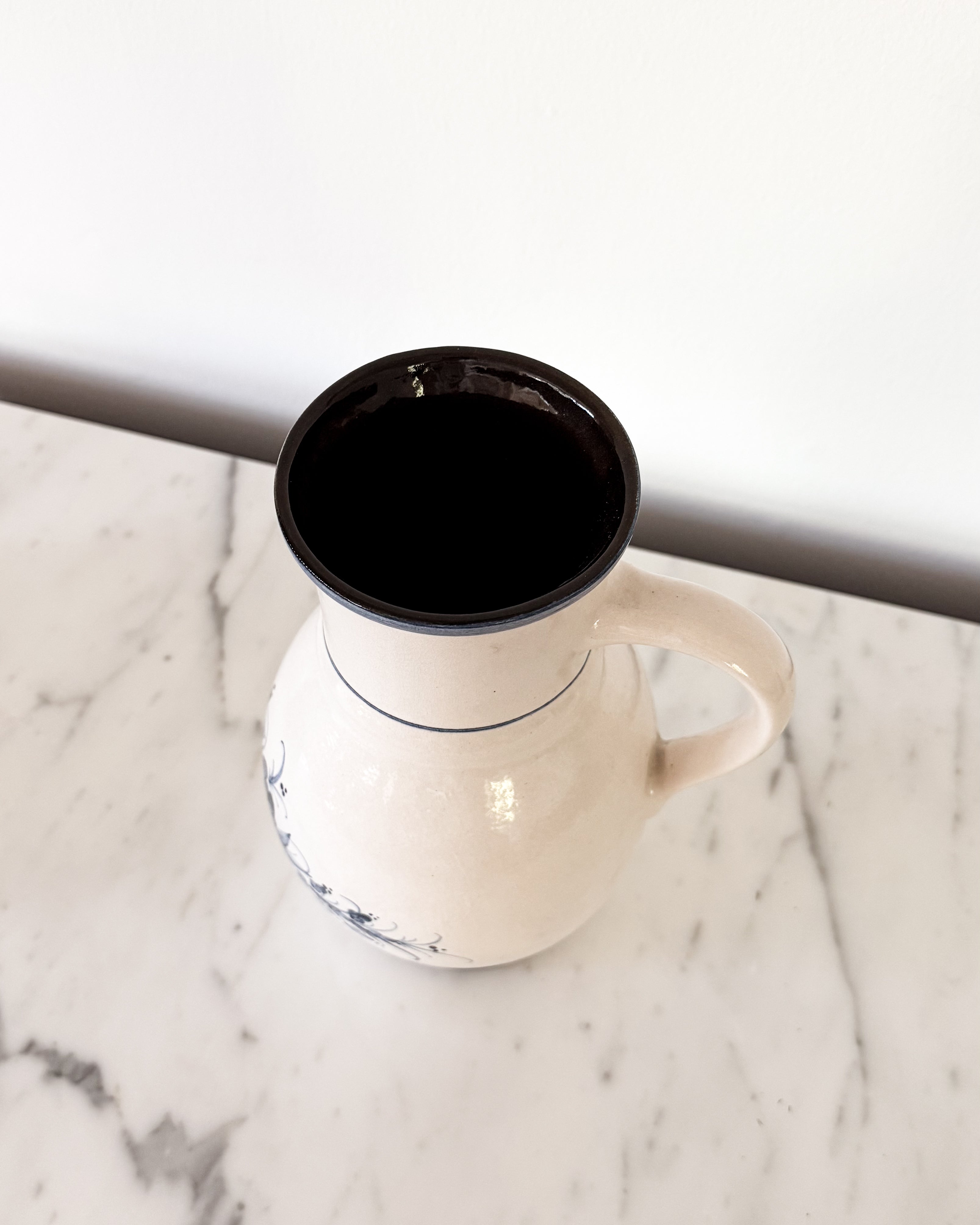 A vintage off-white ceramic pitcher with a prominent rim and handle, featuring subtle painted blue floral patterns on the lower body, set on a white marble surface.