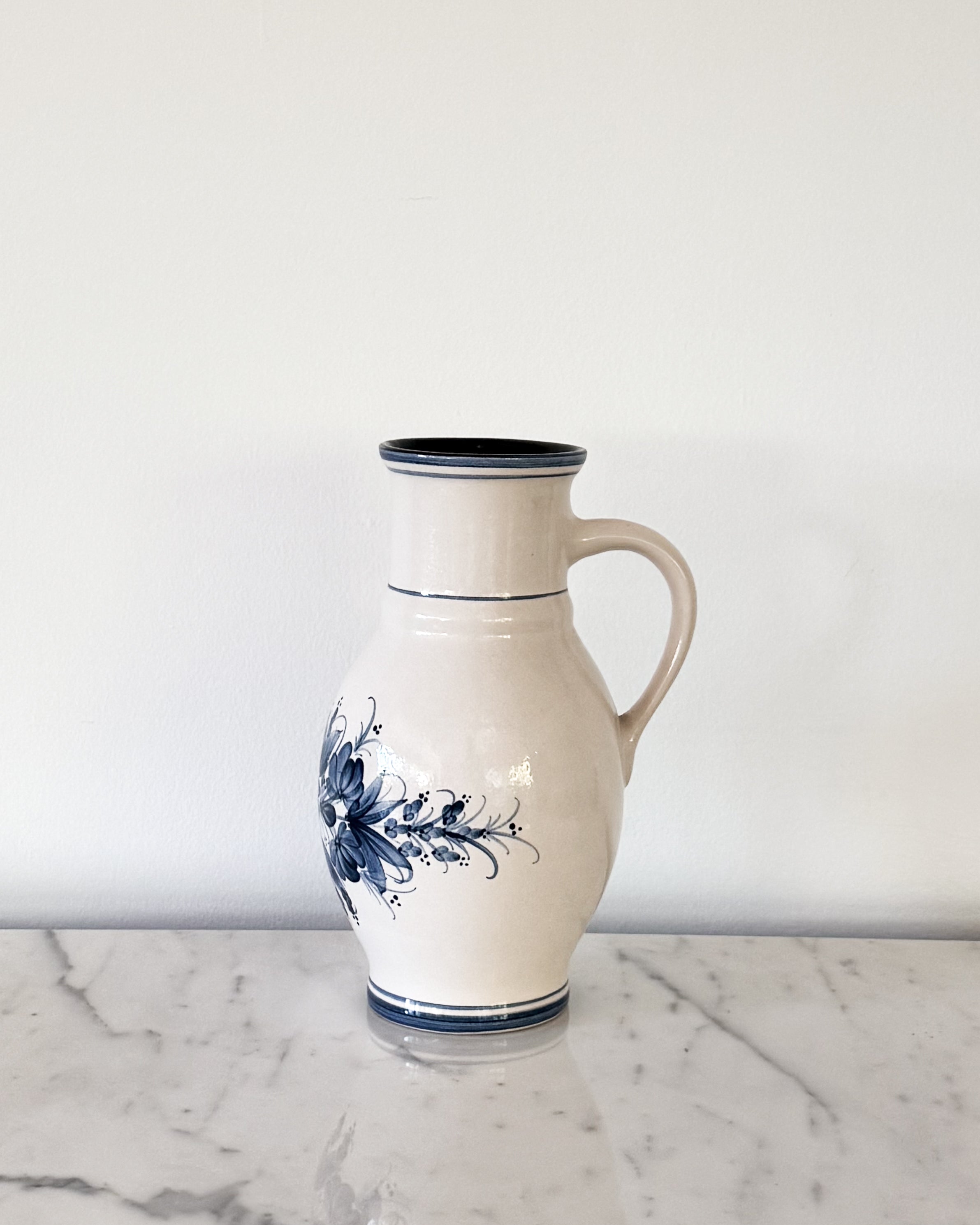 A vintage cream-colored ceramic pitcher with a hand painted blue floral pattern and dark blue accents, set on a white marble surface against a plain white wall.