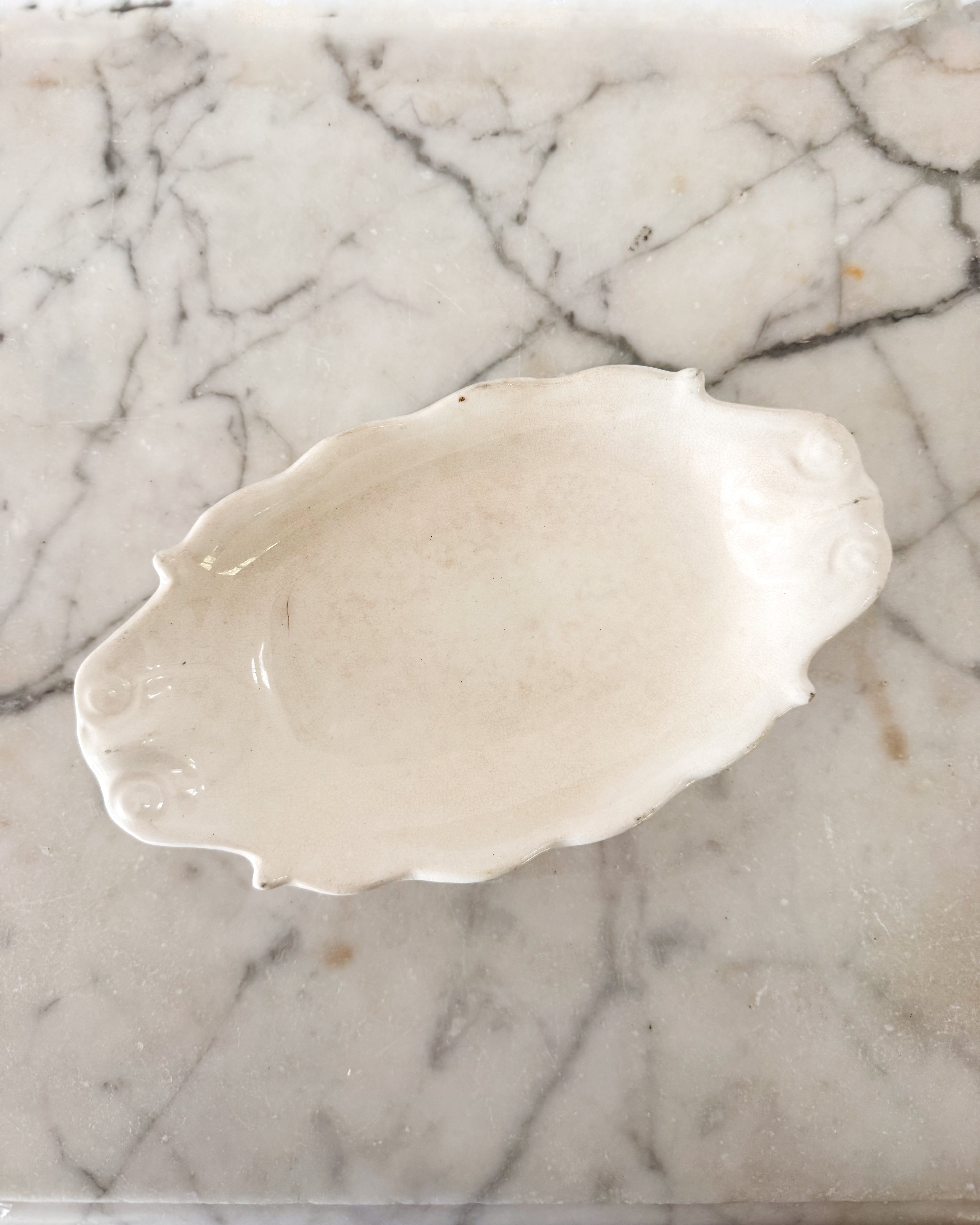A vintage creamy white ironstone spoon rest with a scalloped rim and decorative embossed details, placed on a white marble surface with gray veining.