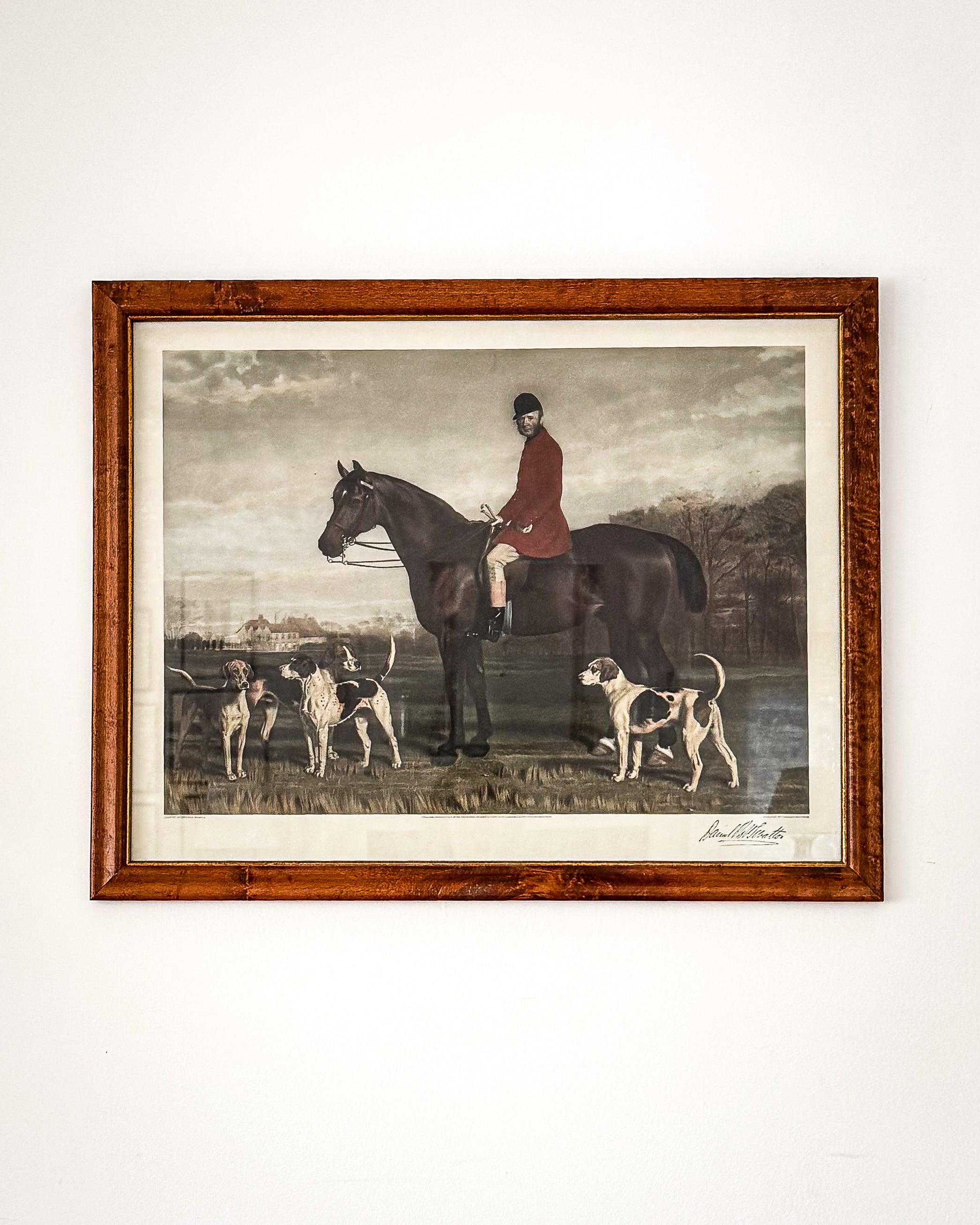 A framed english art print depicting a traditional hunting equestrian scene with a rider on a dark horse and several hounds, mounted on a white wall.
