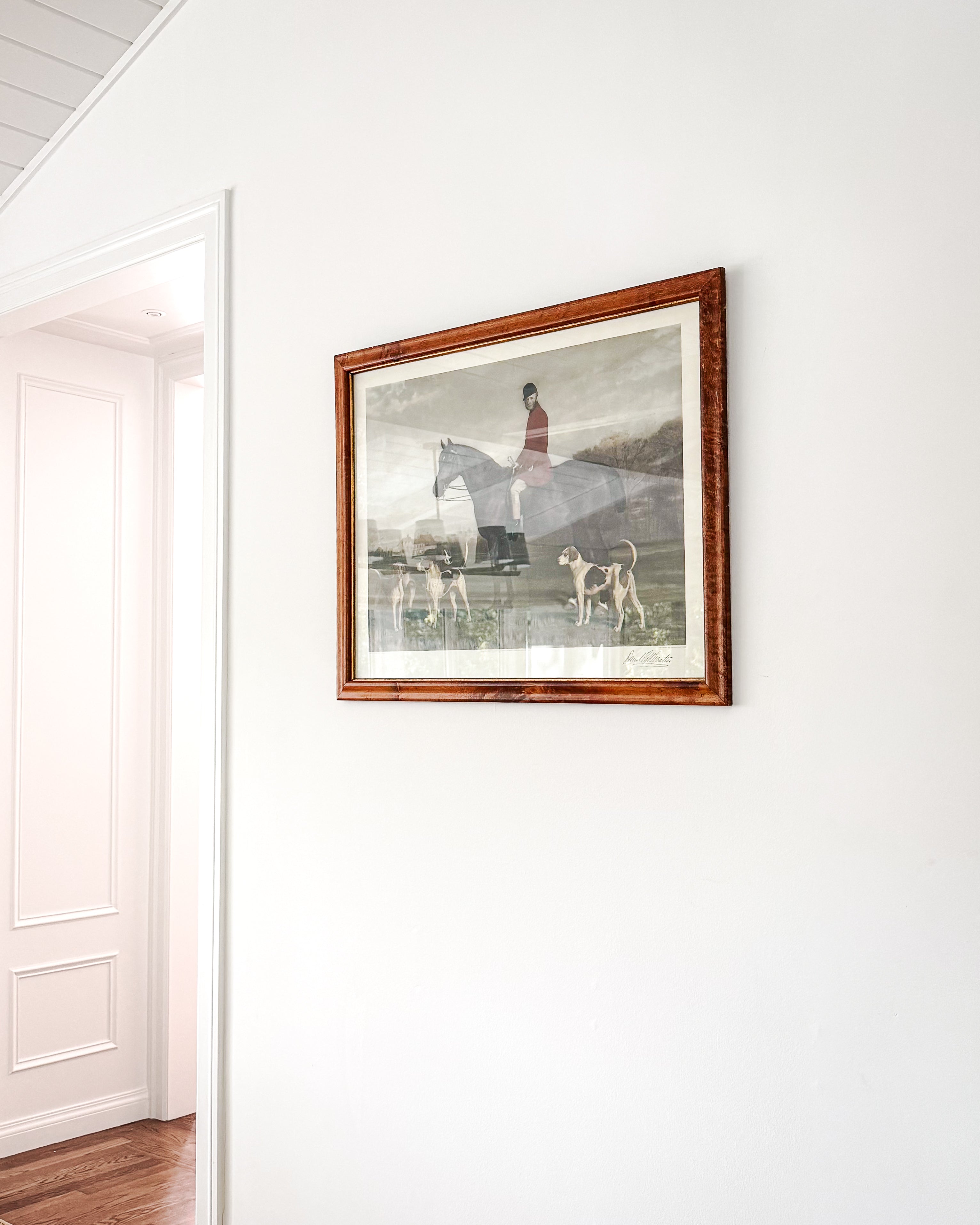 A framed English art print depicting a classic English hunt scene, framed in a burl wood frame. Hung on a white wall in a hallway with a wooden floor visible.