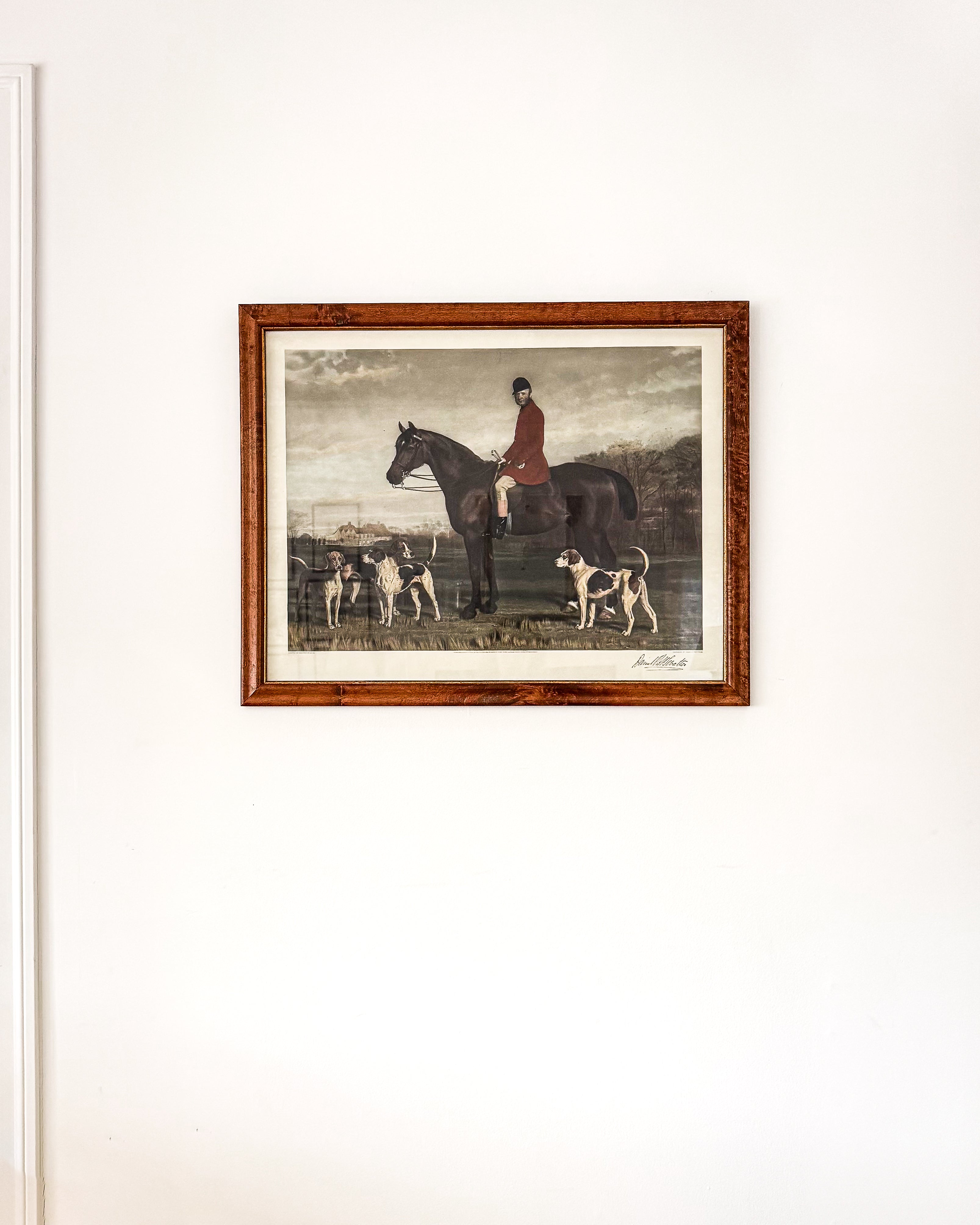 A framed vintage english hunting print in a burl wood frame depicting a rider on horseback with hounds, hung on a plain white wall.