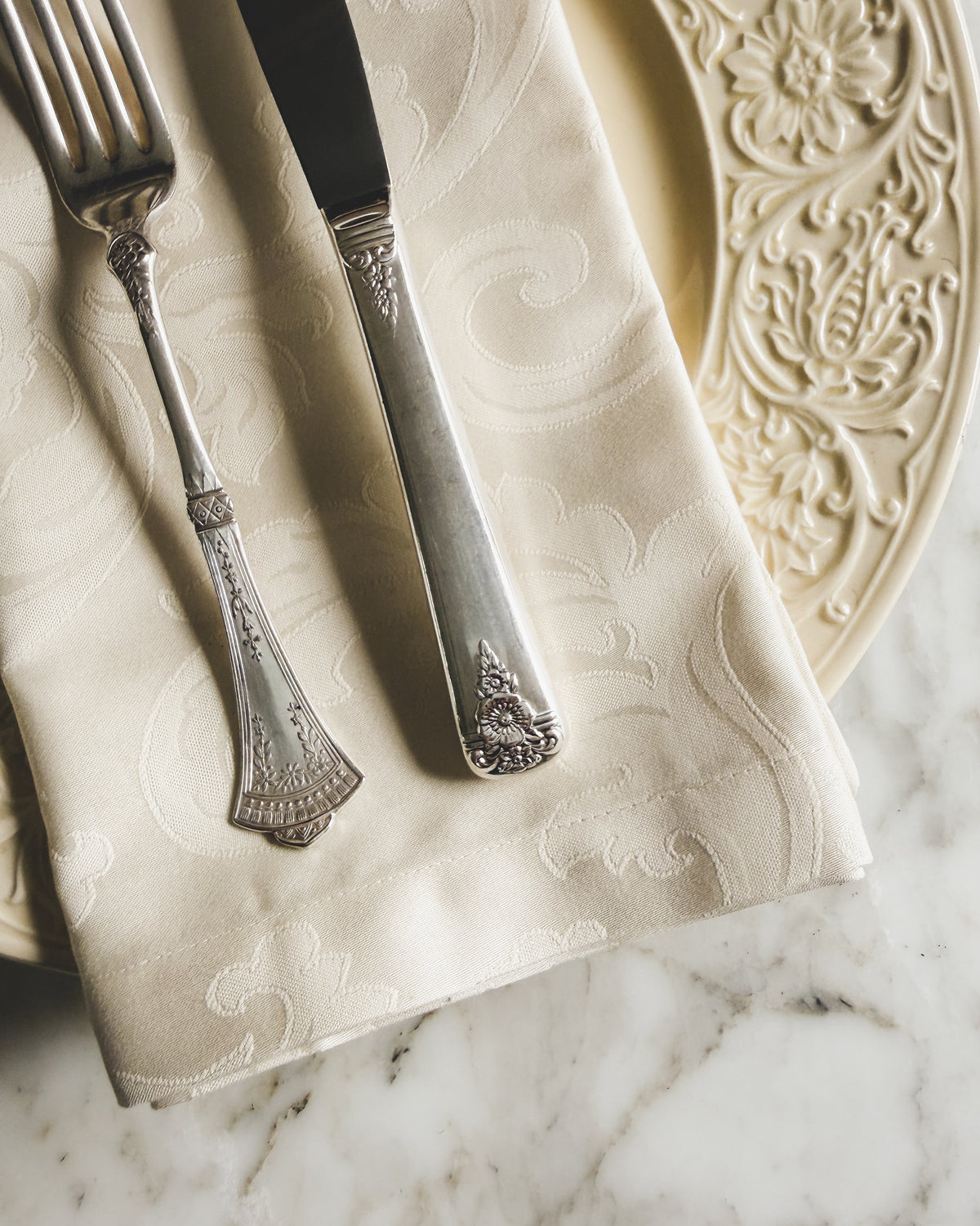 Close-up of mismatched vintage silver plate flatware featuring an ornate floral dinner knife and decorative fork on an ivory damask napkin beside an embossed vintage cream plate.