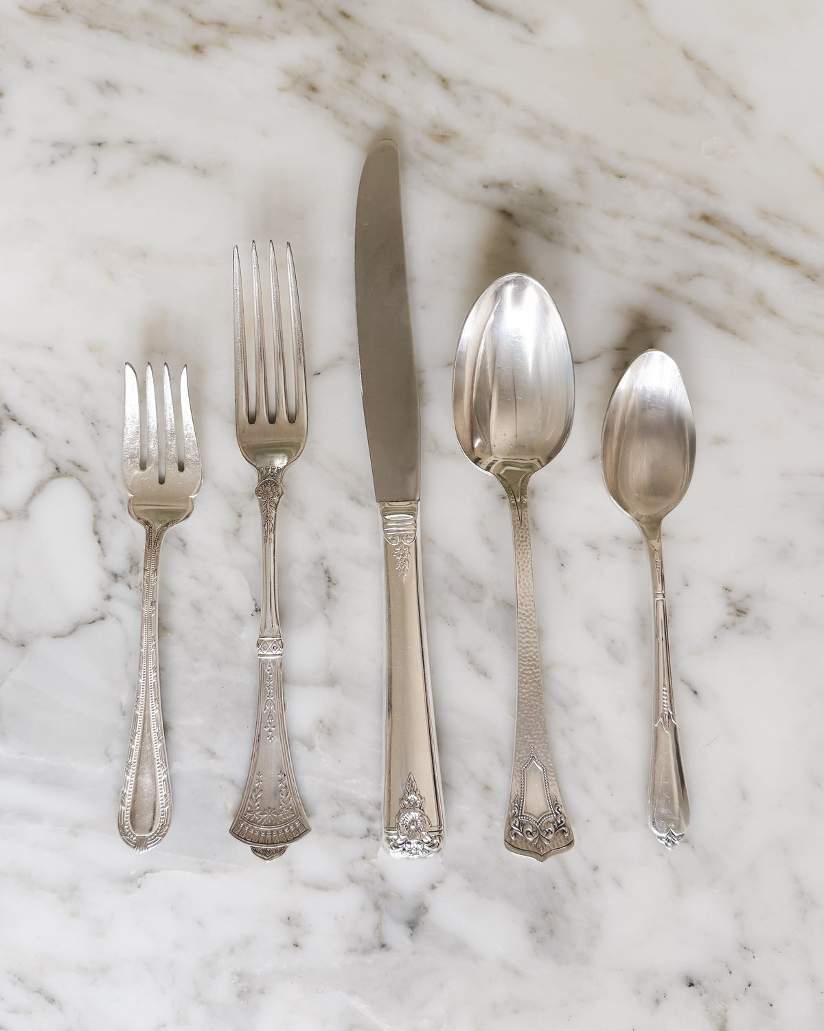 Five-piece place setting of artfully mismatched vintage silver plate flatware, including a dinner fork, salad fork, dinner knife, tablespoon and teaspoon with assorted decorative handles, arranged on white marble.
