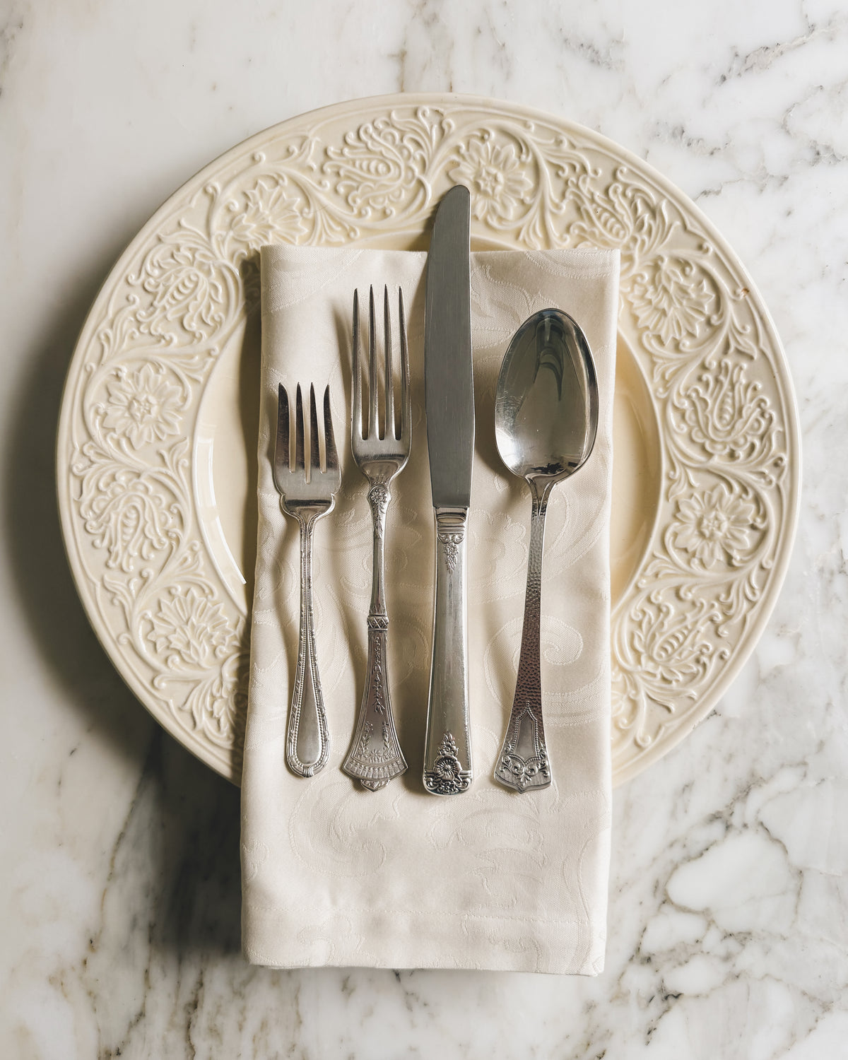 Artfully mismatched vintage silver plate flatware featuring salad fork, dinner fork, a dinner knife and spoon arranged on an ivory Italian damask napkin over an embossed cream dinner plate.