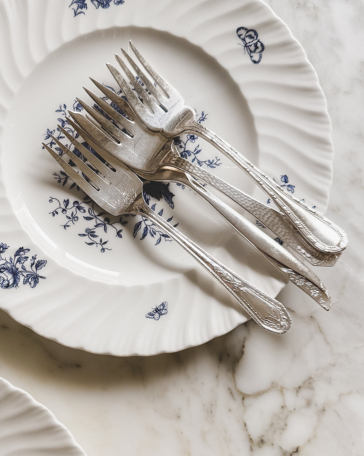 Four artfully mismatched vintage silver plate salad and dessert forks with assorted decorative handles, arranged on a white porcelain plate with delicate blue floral and butterfly motifs.