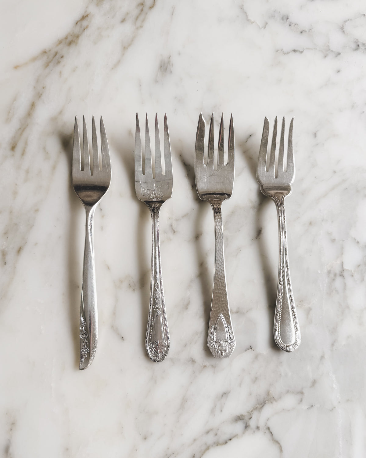 Set of four artfully mismatched vintage silver plate salad and dessert forks, featuring assorted decorative handle patterns, arranged on a white marble surface.
