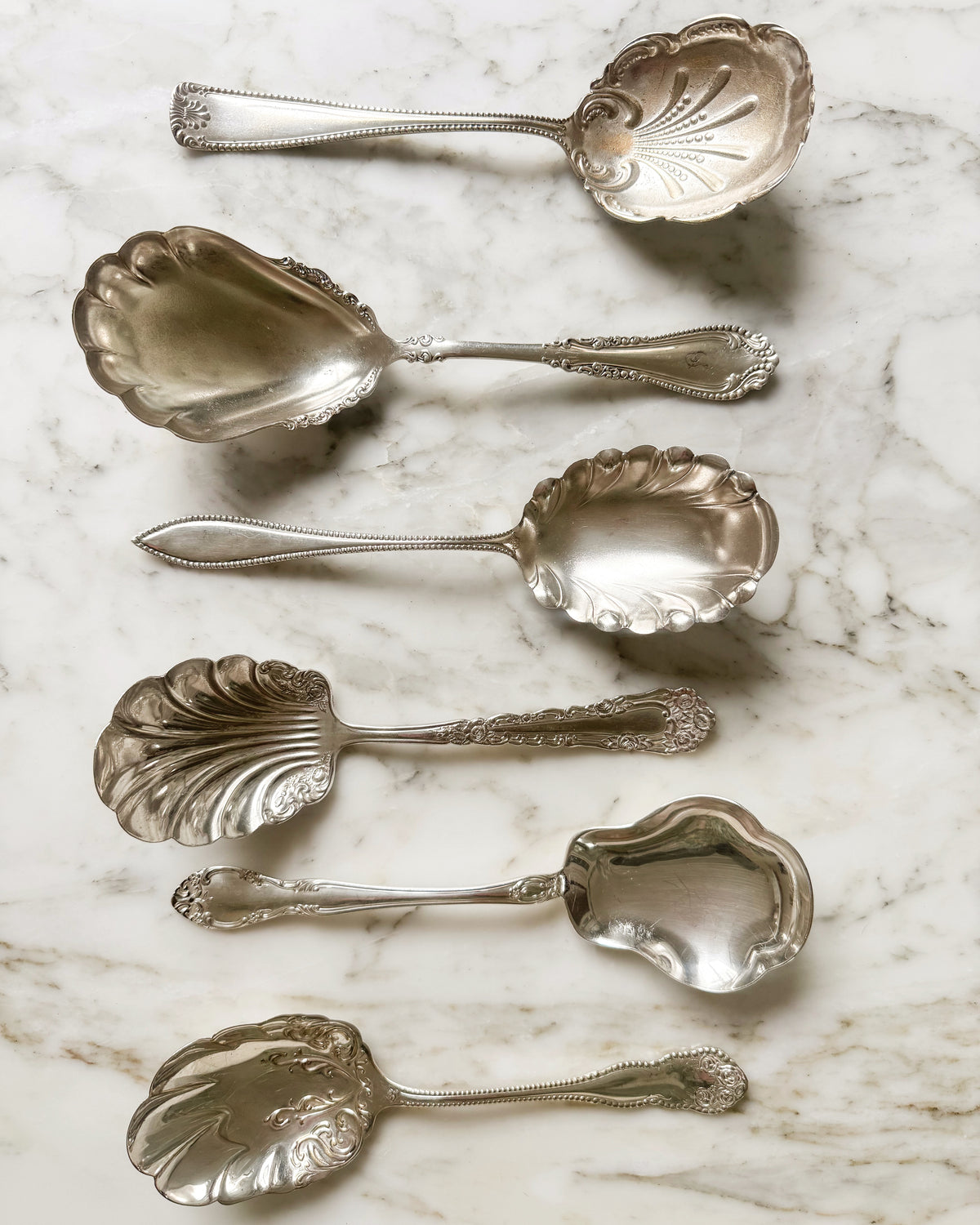 Assorted vintage and antique silver plate shell serving spoons with decorative handles on white marble.