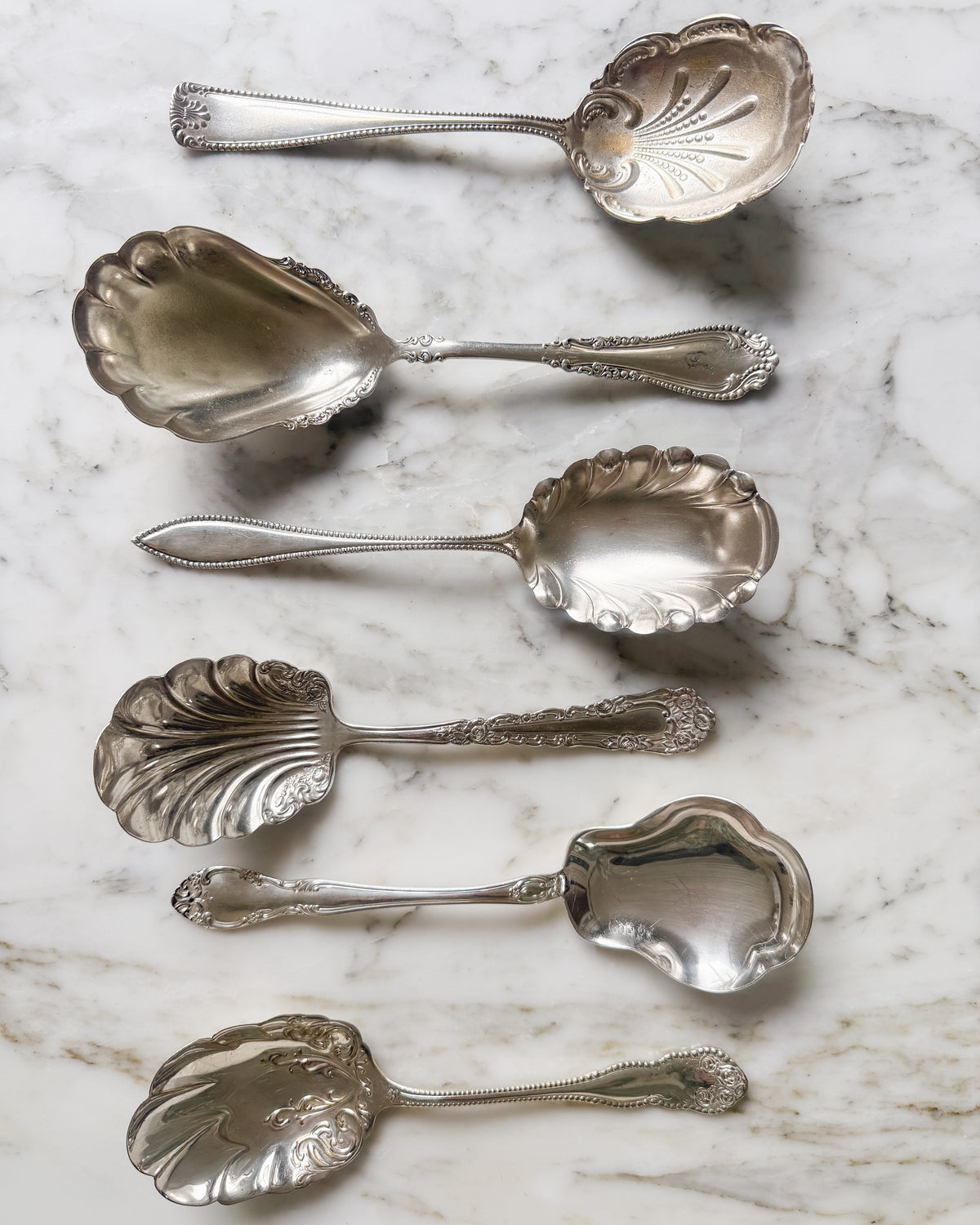 Assorted vintage and antique silver plate shell serving spoons with decorative handles on white marble.